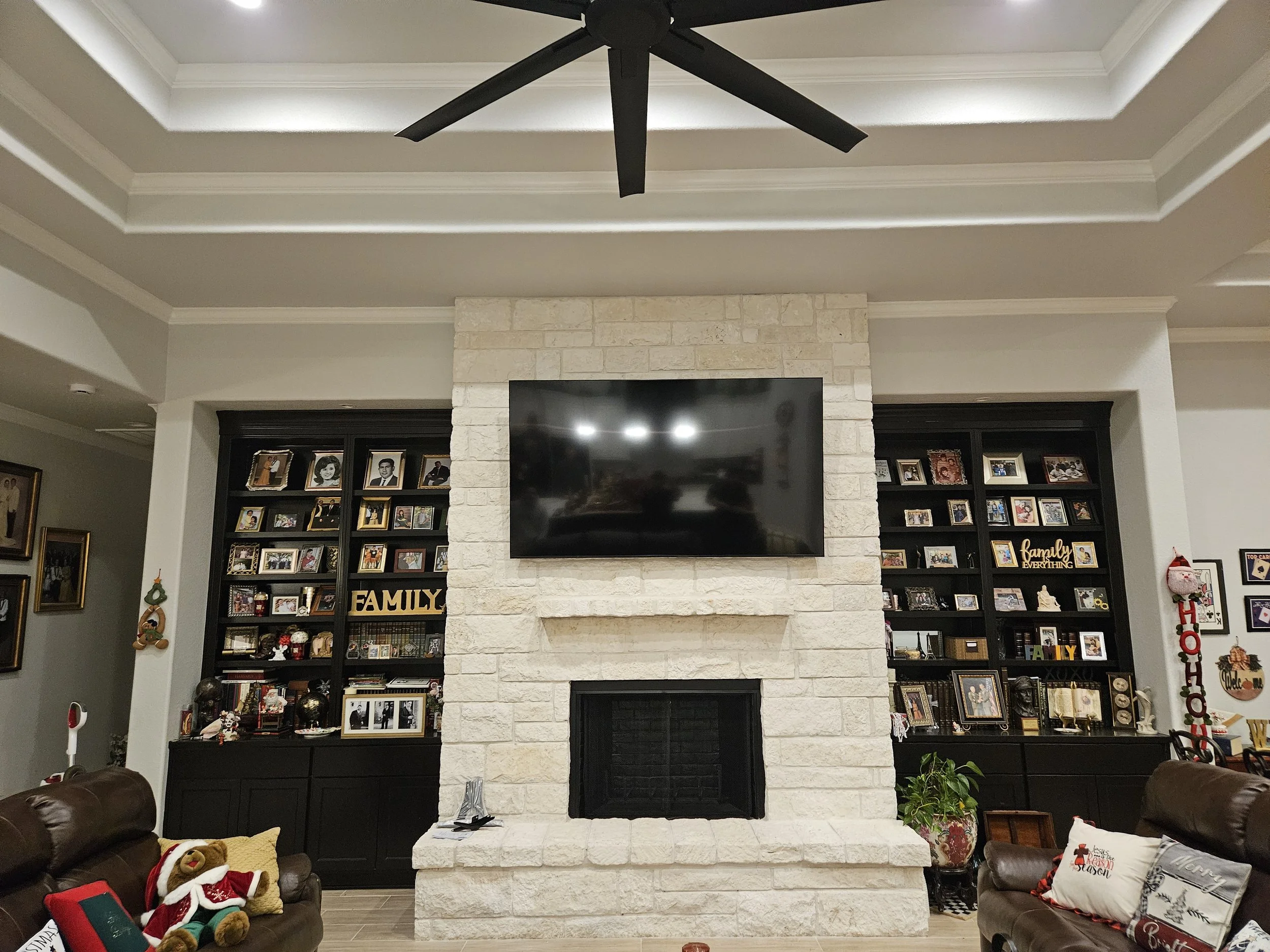 Living room with a white brick fireplace, black TV mounted above, and black bookshelves filled with family photos and decorations. Seating includes brown leather sofas with holiday-themed pillows and a stuffed bear.
