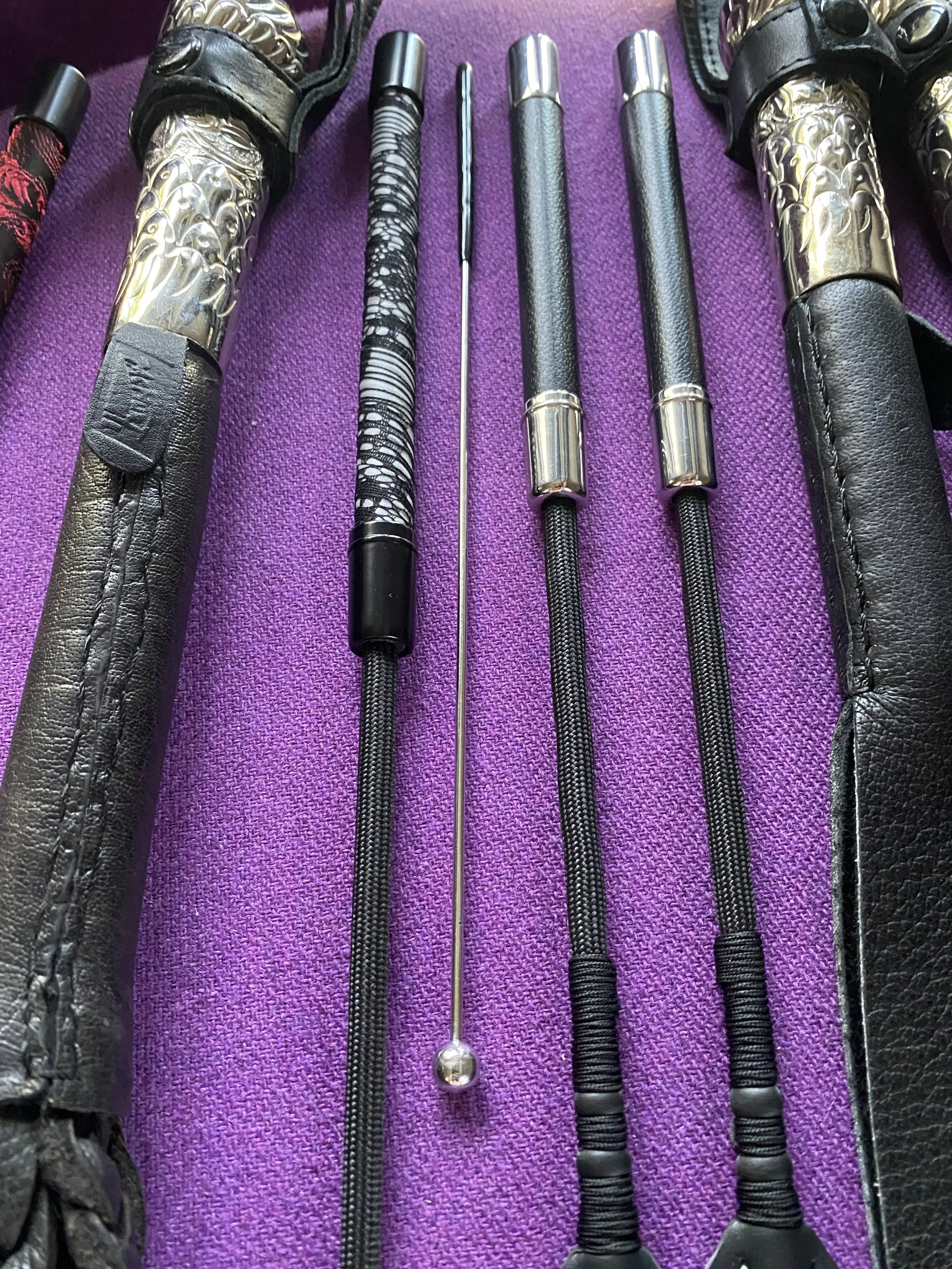 A collection of pool cues with various designs, including pattern and metallic elements, is laid out on a purple fabric surface, with some cues in black leather cases.