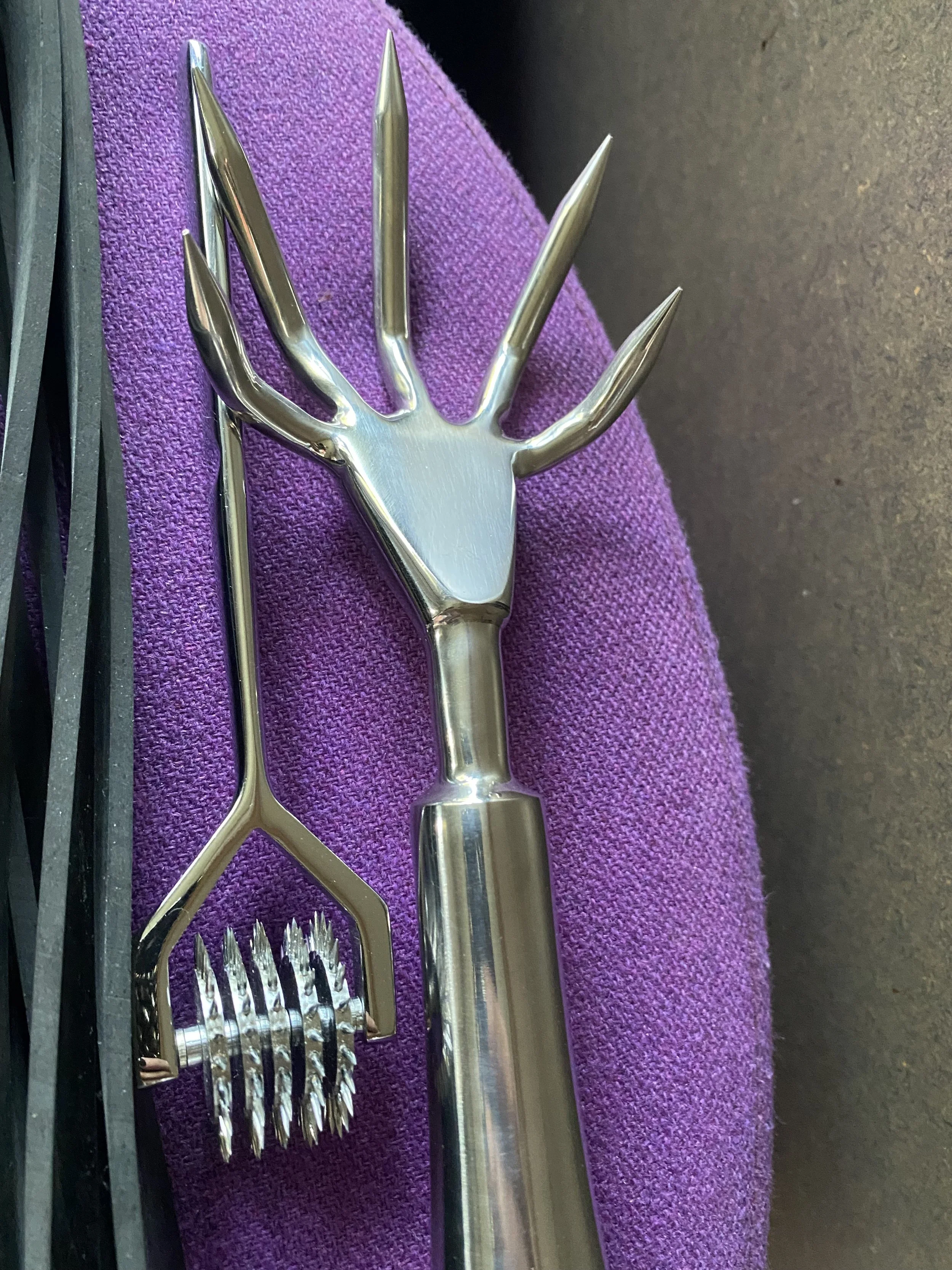 Decorative stainless steel garden tools shaped like a hand with extended fingers and a fork, resting on purple fabric and black surface.