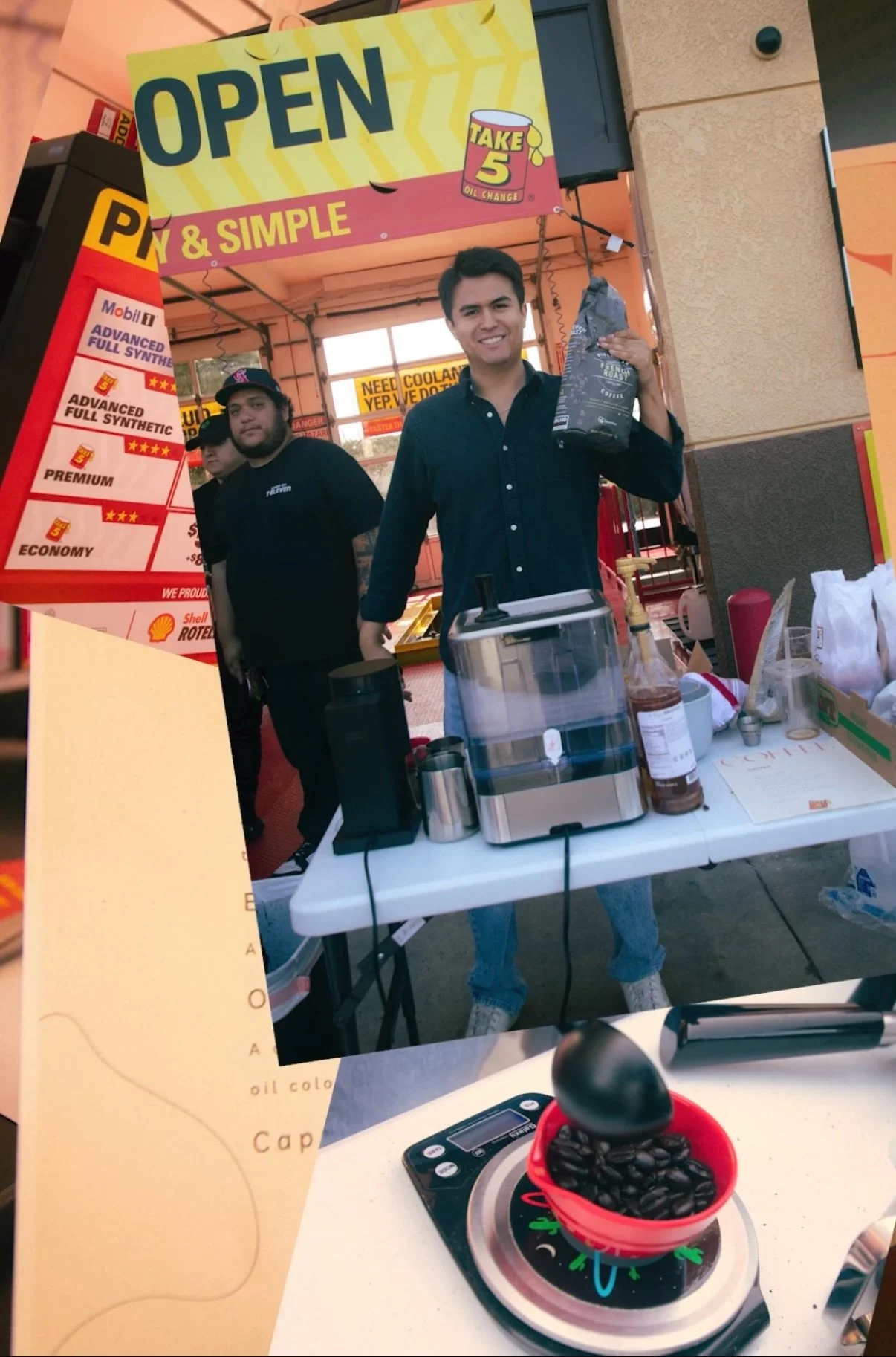 Young man in a black shirt holding a bag of coffee beans, standing at a coffee stand with a table that has a coffee grinder and syrup bottles, with two other men behind him. There is a large yellow and pink sign indicating the stand is open.