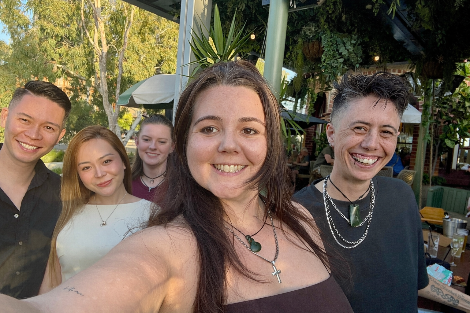 Group of five young adults smiling for a selfie at an outdoor gathering, with trees and patio furniture in the background during daytime