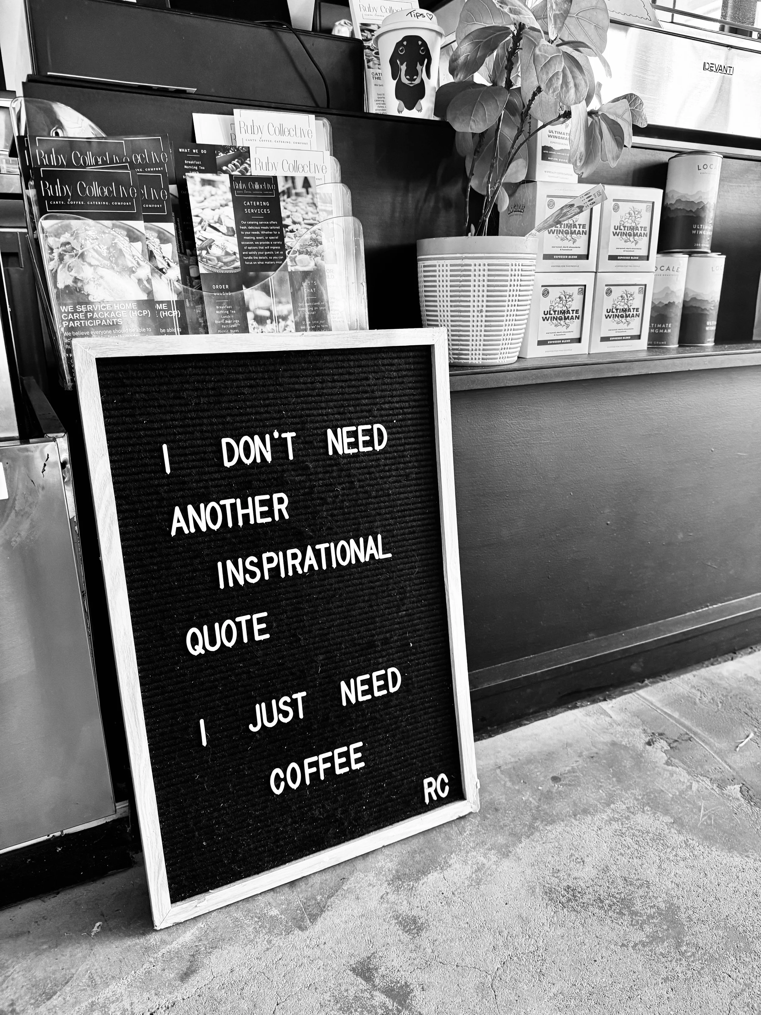 A black felt letter board sign with white letters placed on the floor in front of a counter. The sign reads: 'I DON'T NEED ANOTHER INSPIRATIONAL QUOTE I JUST NEED COFFEE RC'. Behind the sign, there is a counter with magazines, brochures, a potted plant, and some boxed items. There are also disposable cups and other promotional materials on the counter.