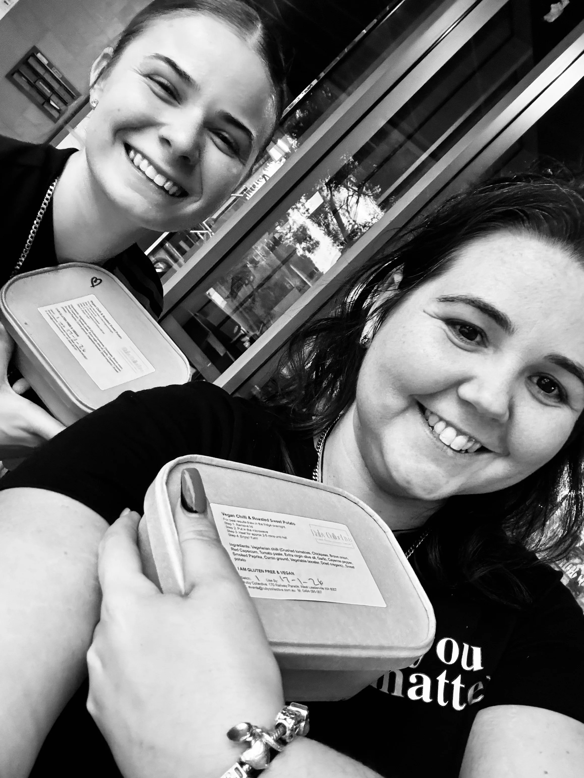 Two women smiling and holding meal containers, standing outside a building with large windows.