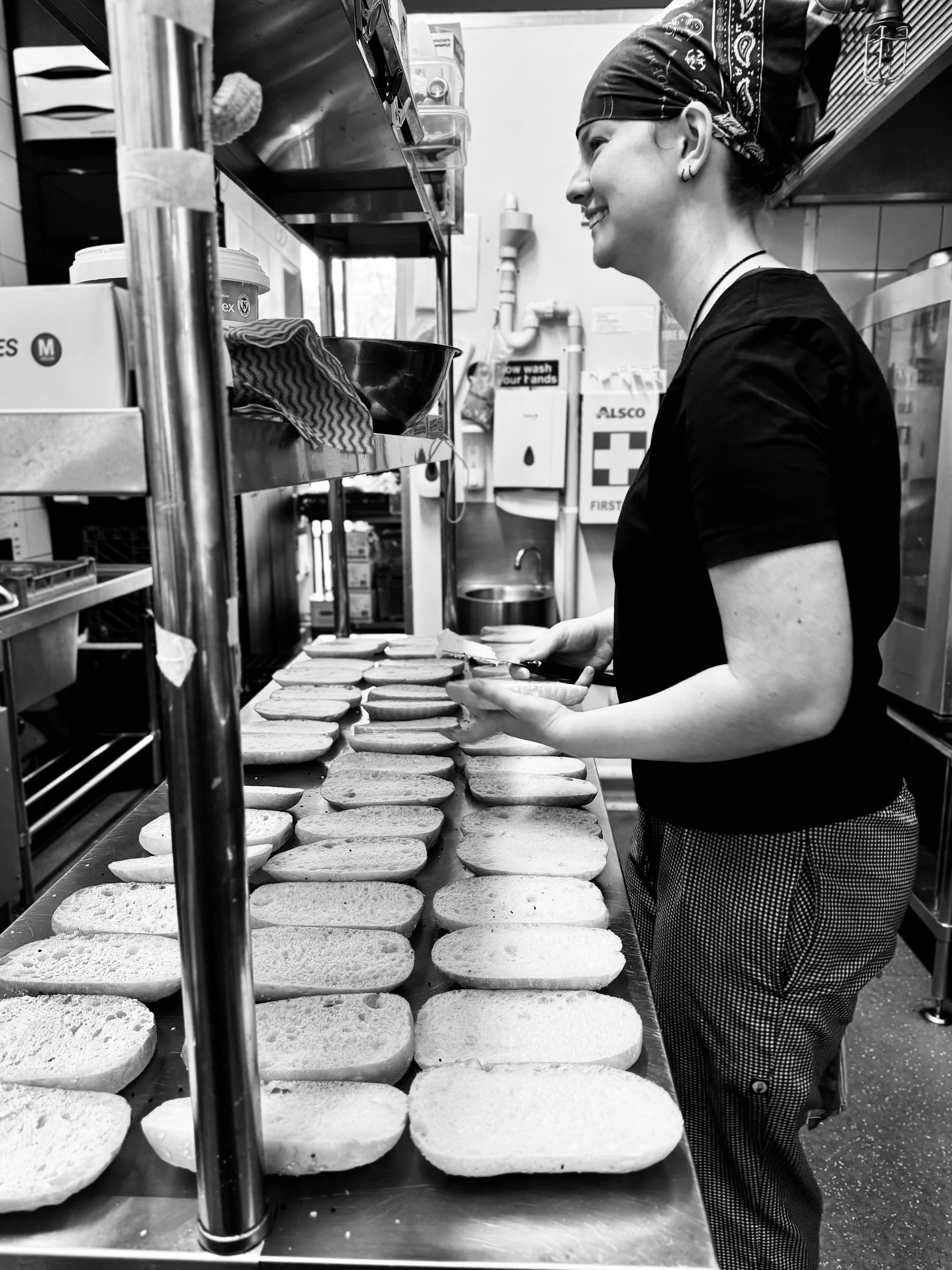 A woman in a bandana and black shirt preparing sandwiches on a stainless steel counter in a commercial kitchen.