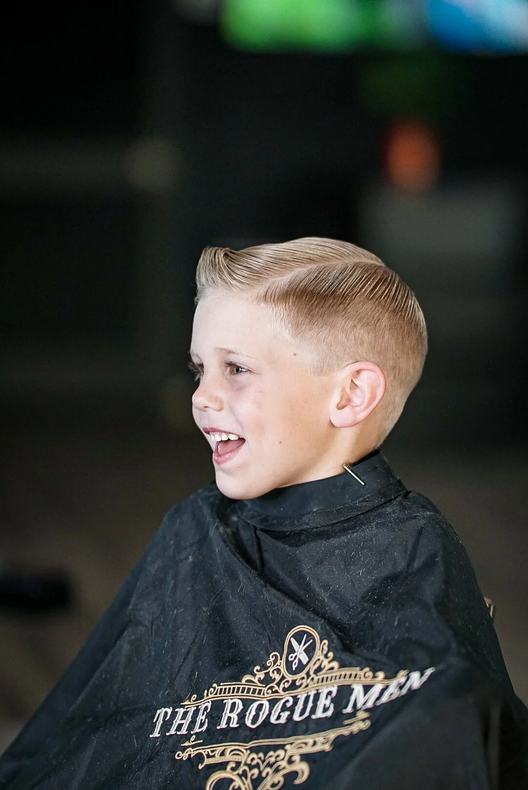 A young boy with blonde hair smiling and wearing a black barber cape at a barbershop.