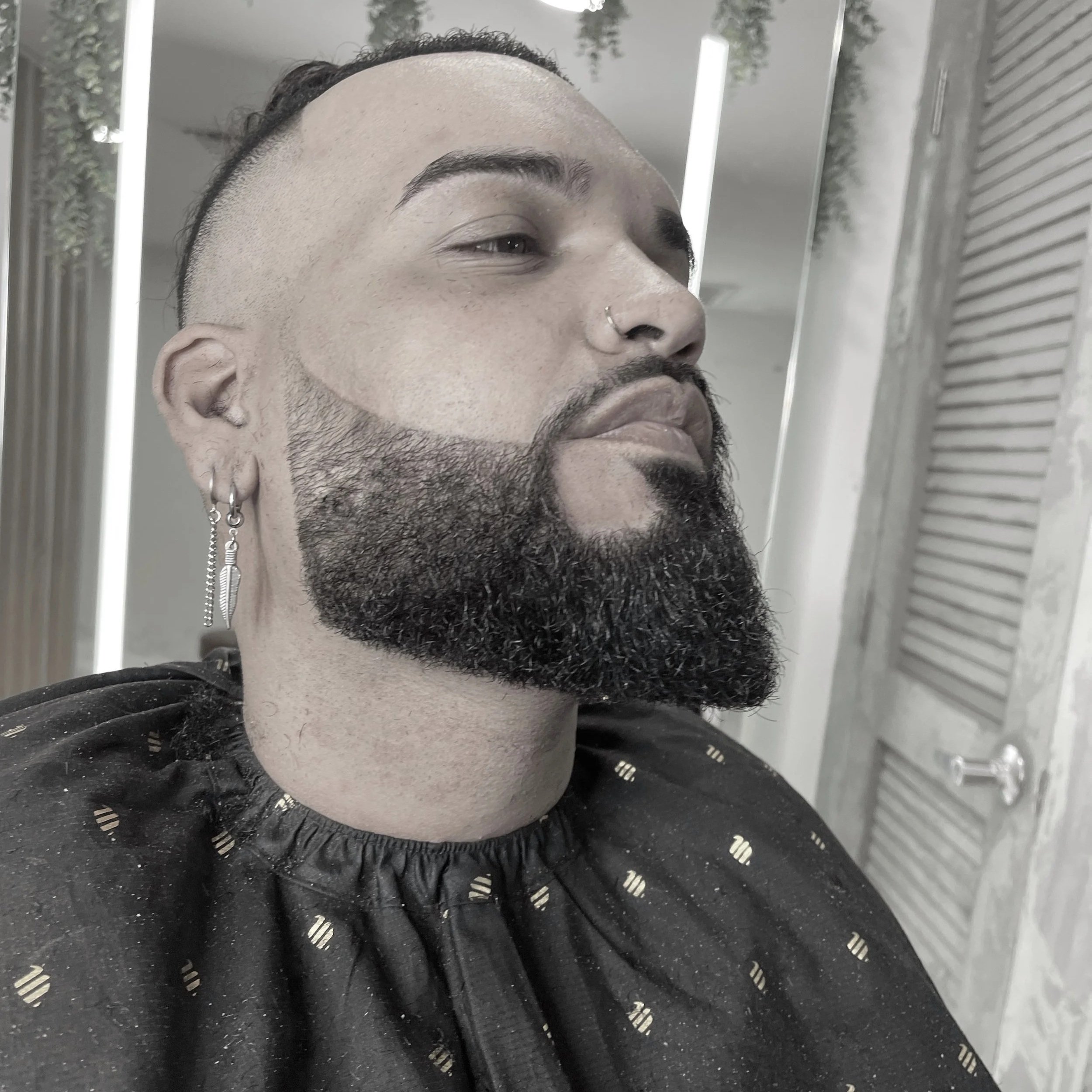 A man with a full beard and pierced ears, sitting in a salon chair, wearing a black cape, and looking slightly to the side with a serious expression. The background includes a mirror, hanging plants, and a door.