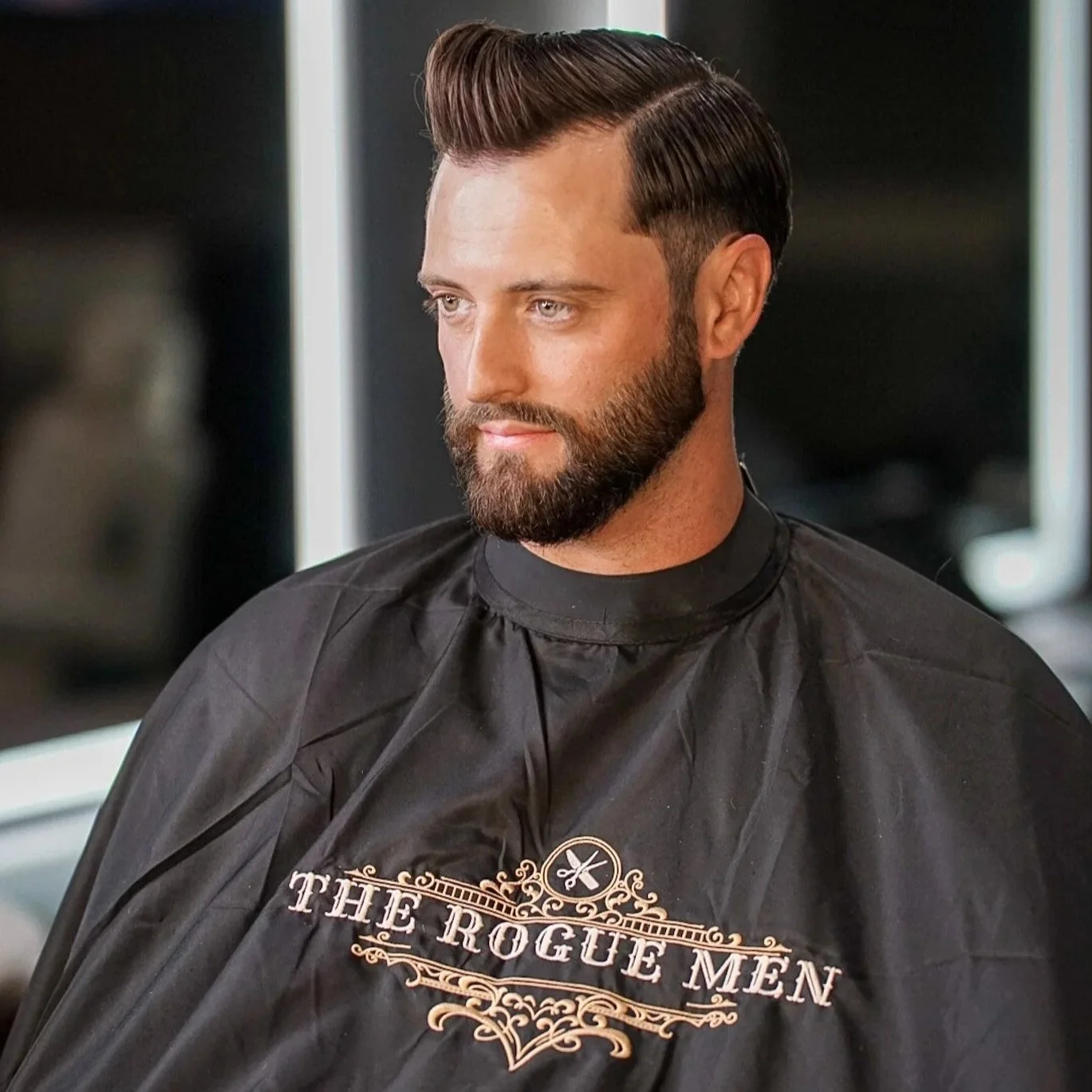 A man with styled dark hair and beard is sitting in a barber's chair wearing a black barber cape with the logo 'The Rogue Men.'