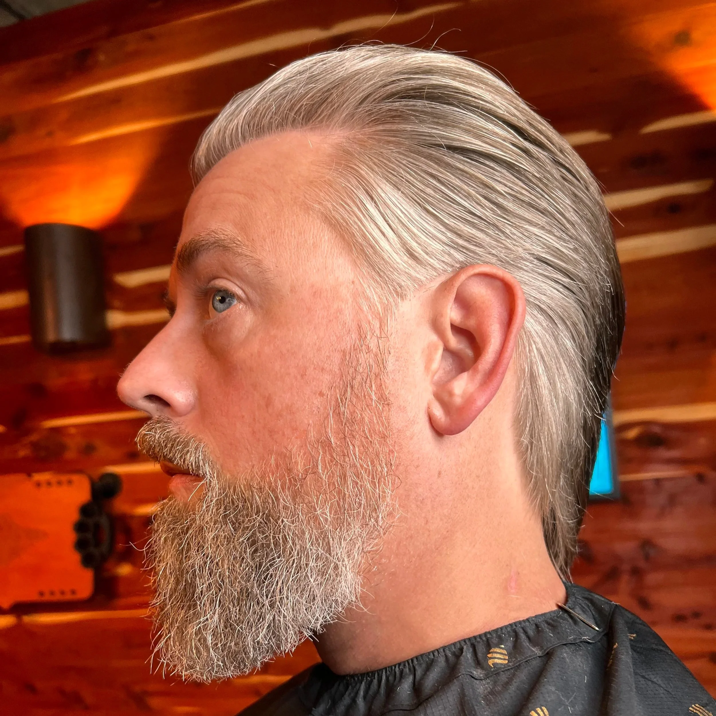 Side profile of a middle-aged man with gray hair and a full beard, sitting in a barber's chair with a black cape. The background features warm wooden paneling and wall-mounted lights.