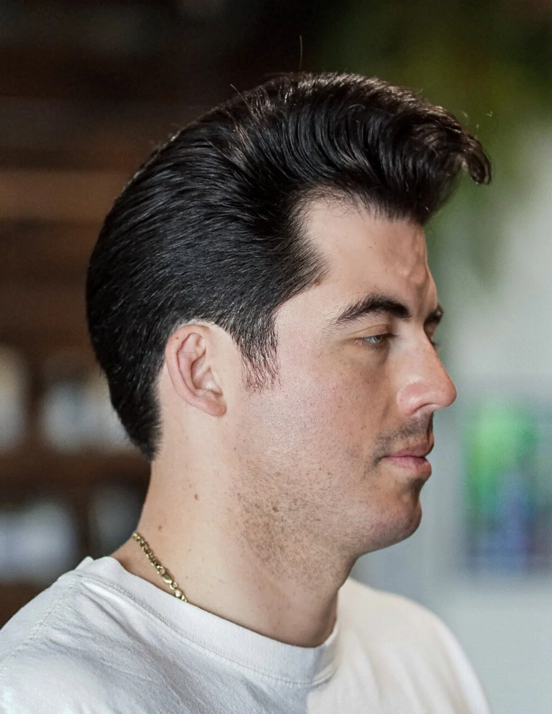 Side profile of a young man with dark, slicked-back hair, wearing a white shirt and a gold chain necklace.