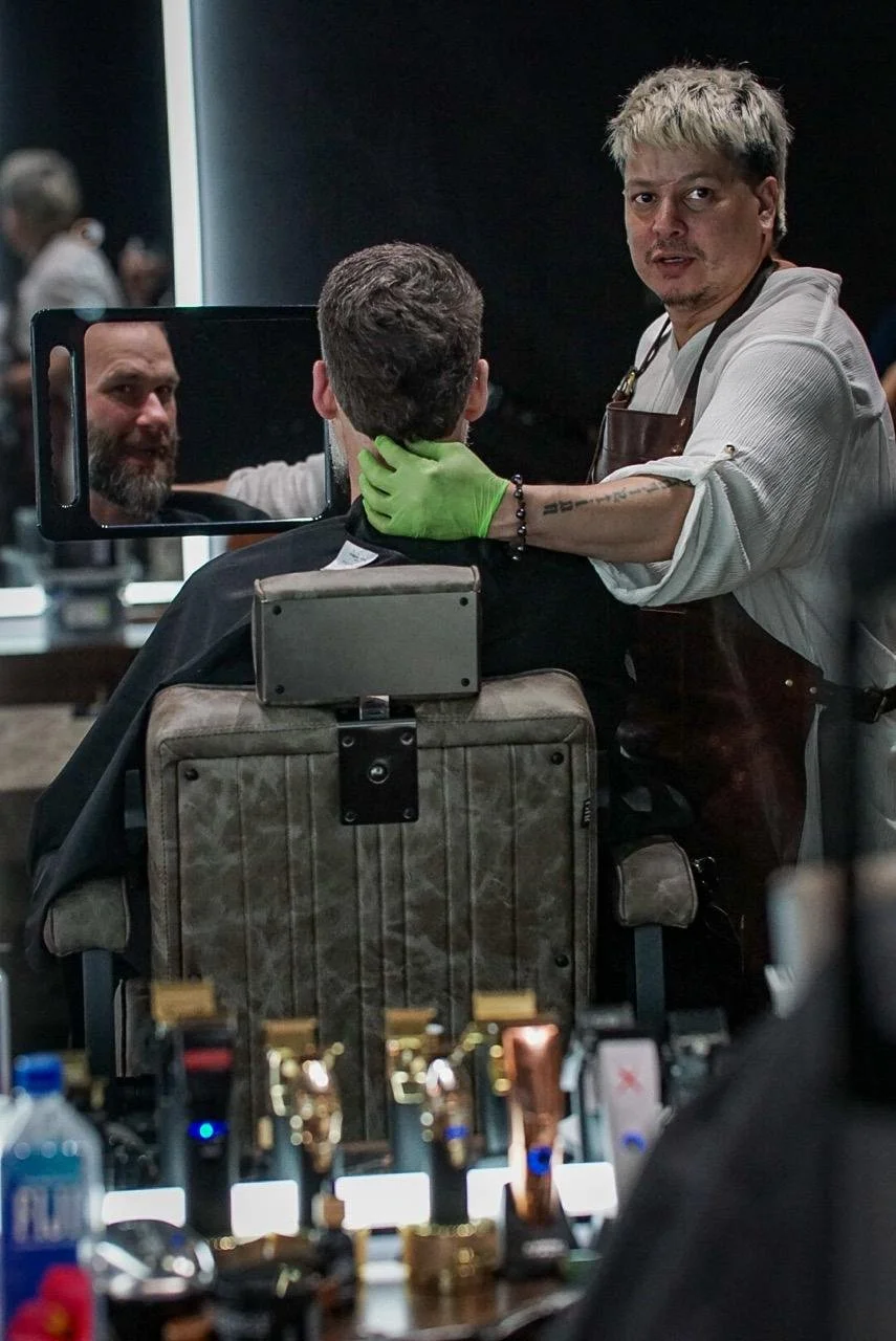 A hairstylist wearing an apron and green gloves cuts a customer's hair, seen from behind, with a mirror reflecting the customer's face. The scene takes place in a salon with hair styling tools on the counter in the foreground.