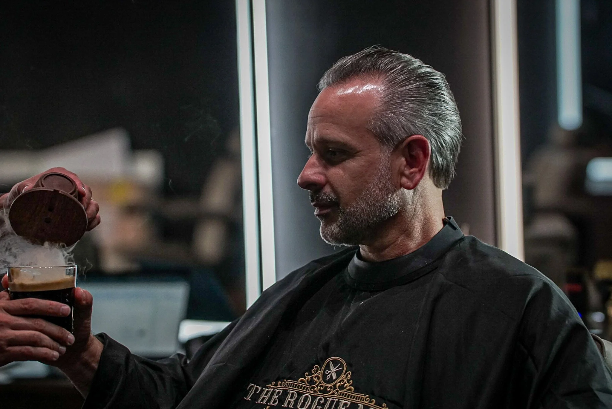 A man with gray hair and a beard getting a cold drink poured into a glass, wearing a black barber cape with logo, in a barbershop or salon setting.