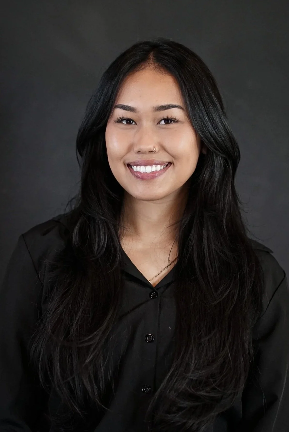 Portrait of a young woman with long black hair, wearing a black shirt and smiling, against a dark background.