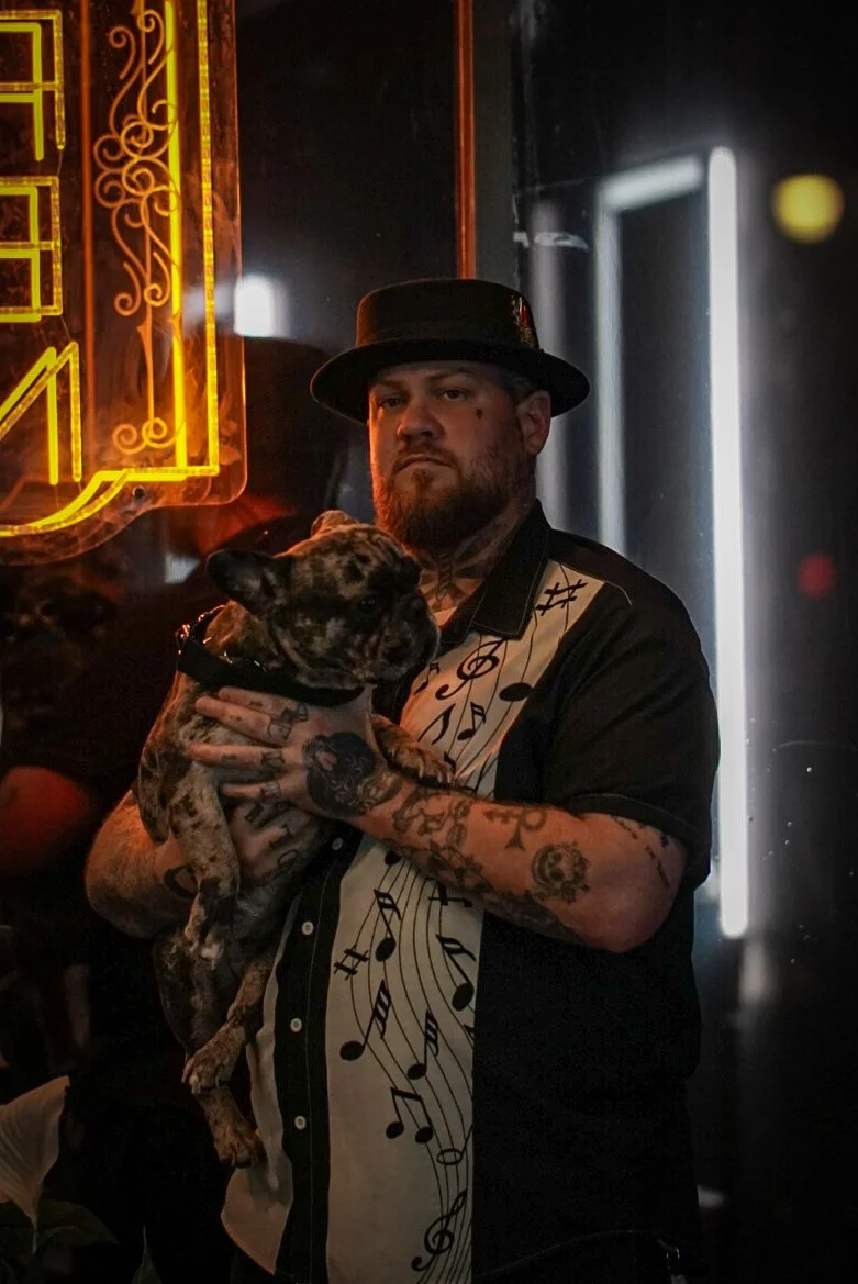 A man with a beard, tattoos, and a black hat holding a brindle French bulldog, standing in a dimly lit setting with a neon sign and dark walls.