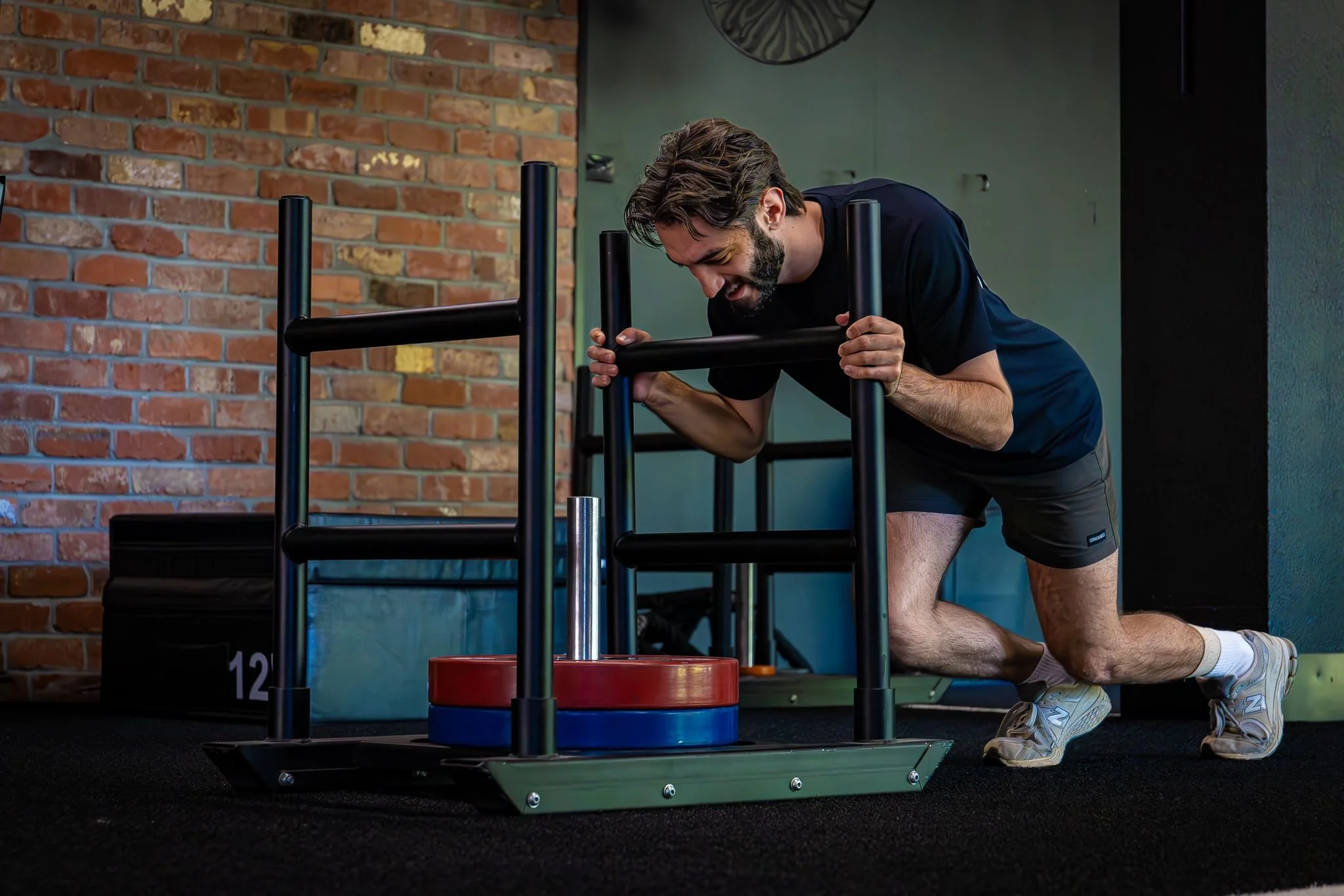 Personal trainer performing a push in gym with sled weights on black carpet floor, brick wall background.