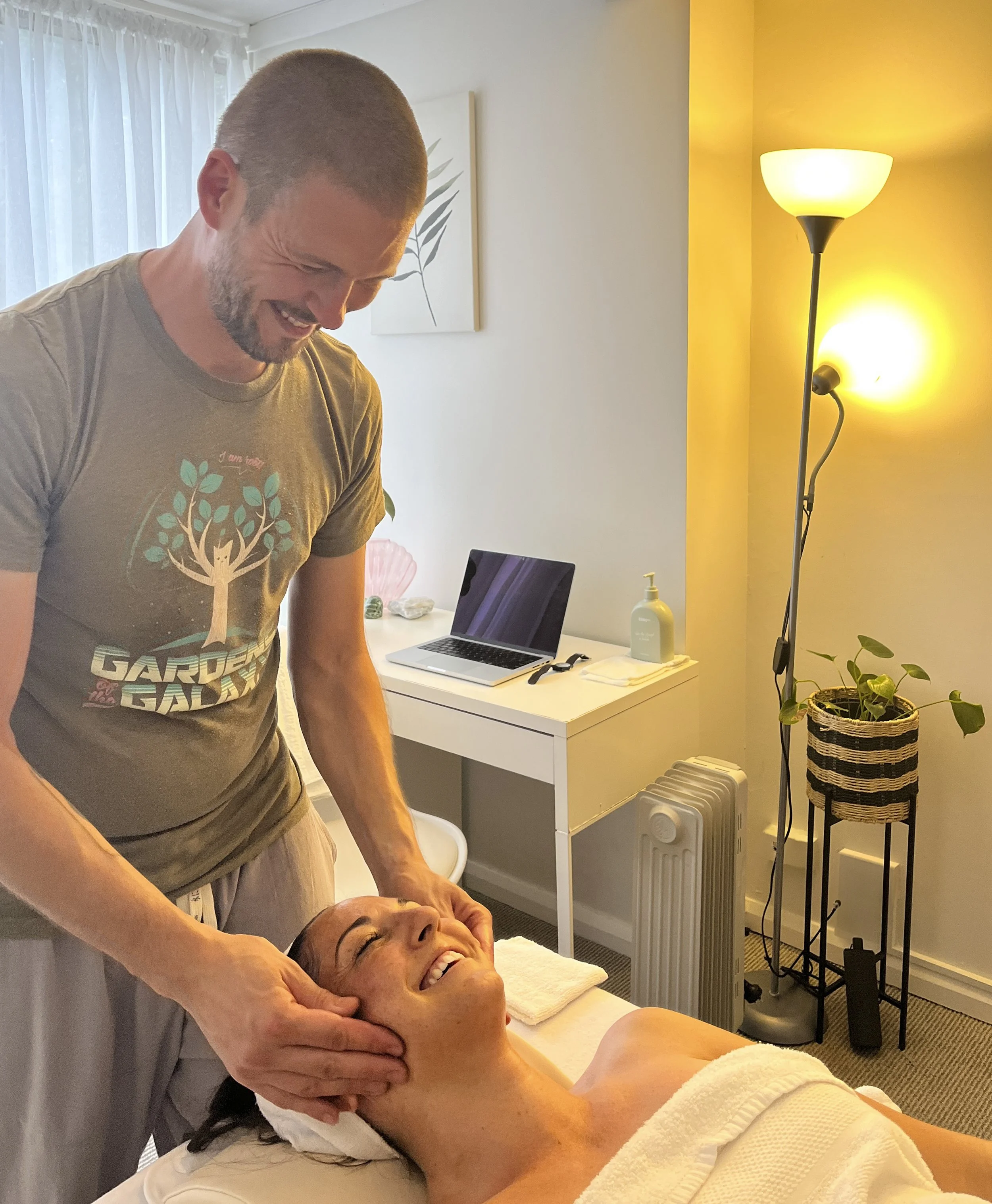 A person receiving a facial massage in a relaxing spa environment, with a therapist smiling. The room is softly lit, featuring a lamp, a small table, a plant, and bottled oil. The client appears relaxed and comfortable.