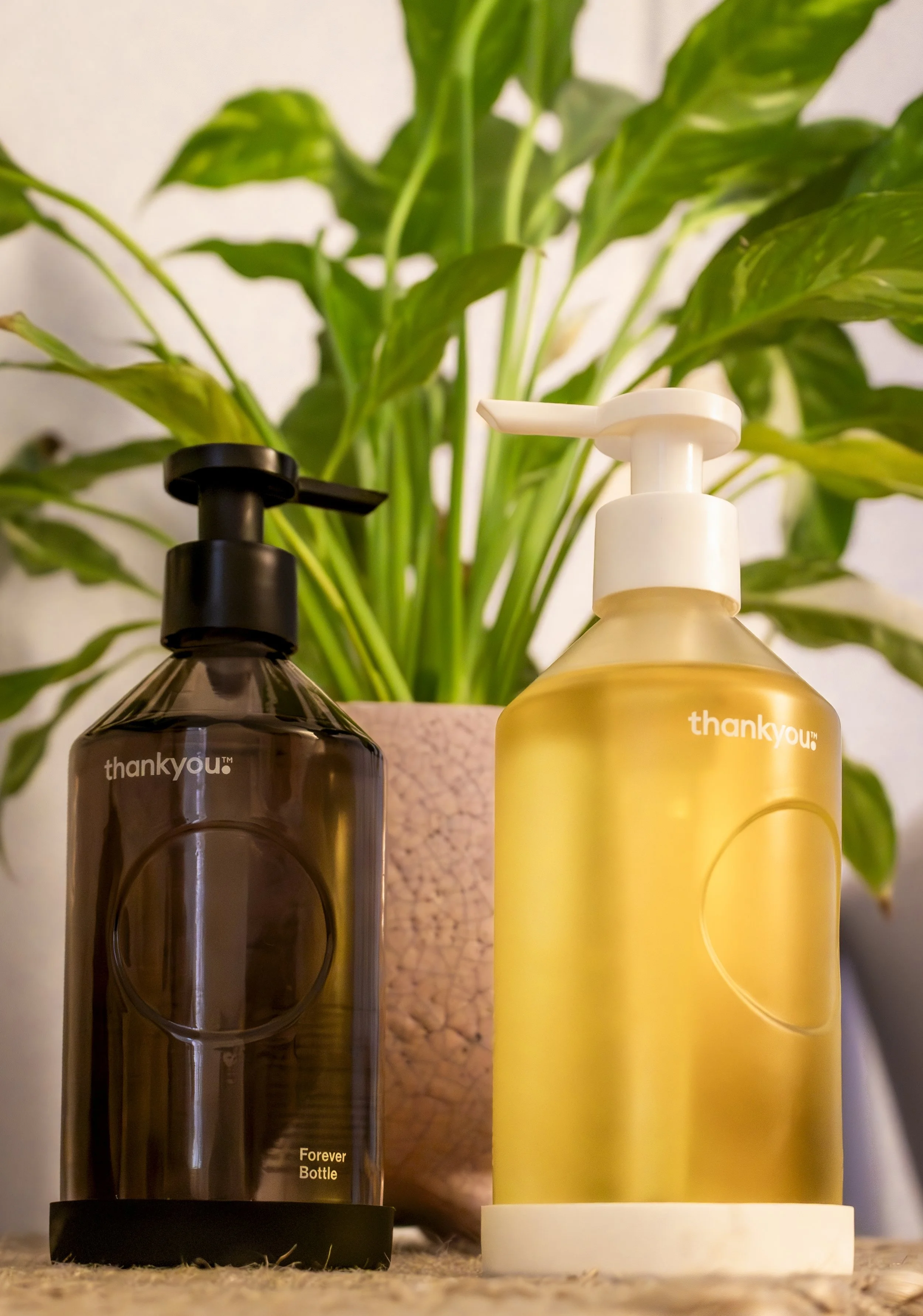 Two massage oil dispensers, one brown and one white, labelled 'thank you', placed on a surface with a green potted plant in the background.