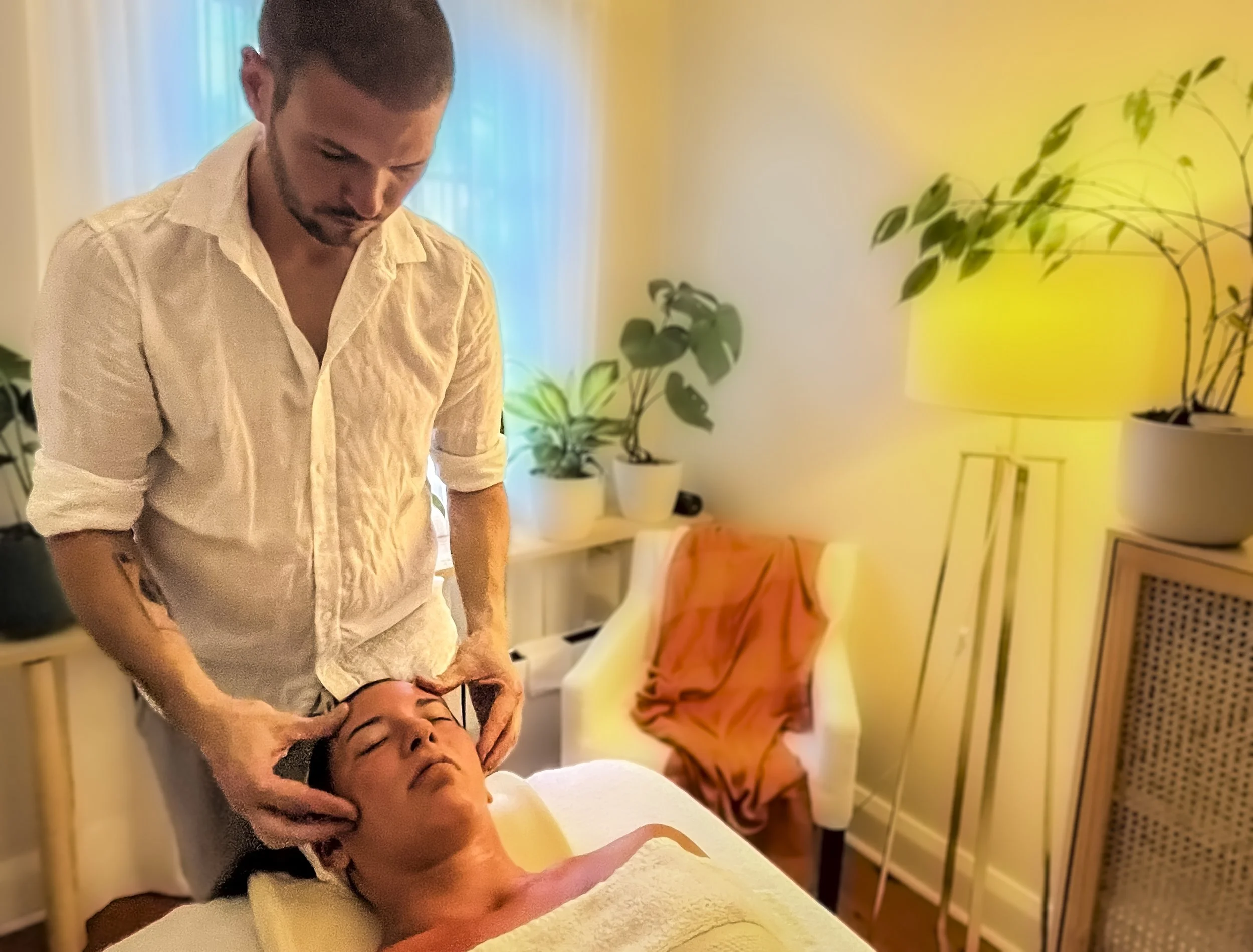 A person receiving a head massage from a massage therapist in a serene room with plants and soft lighting.