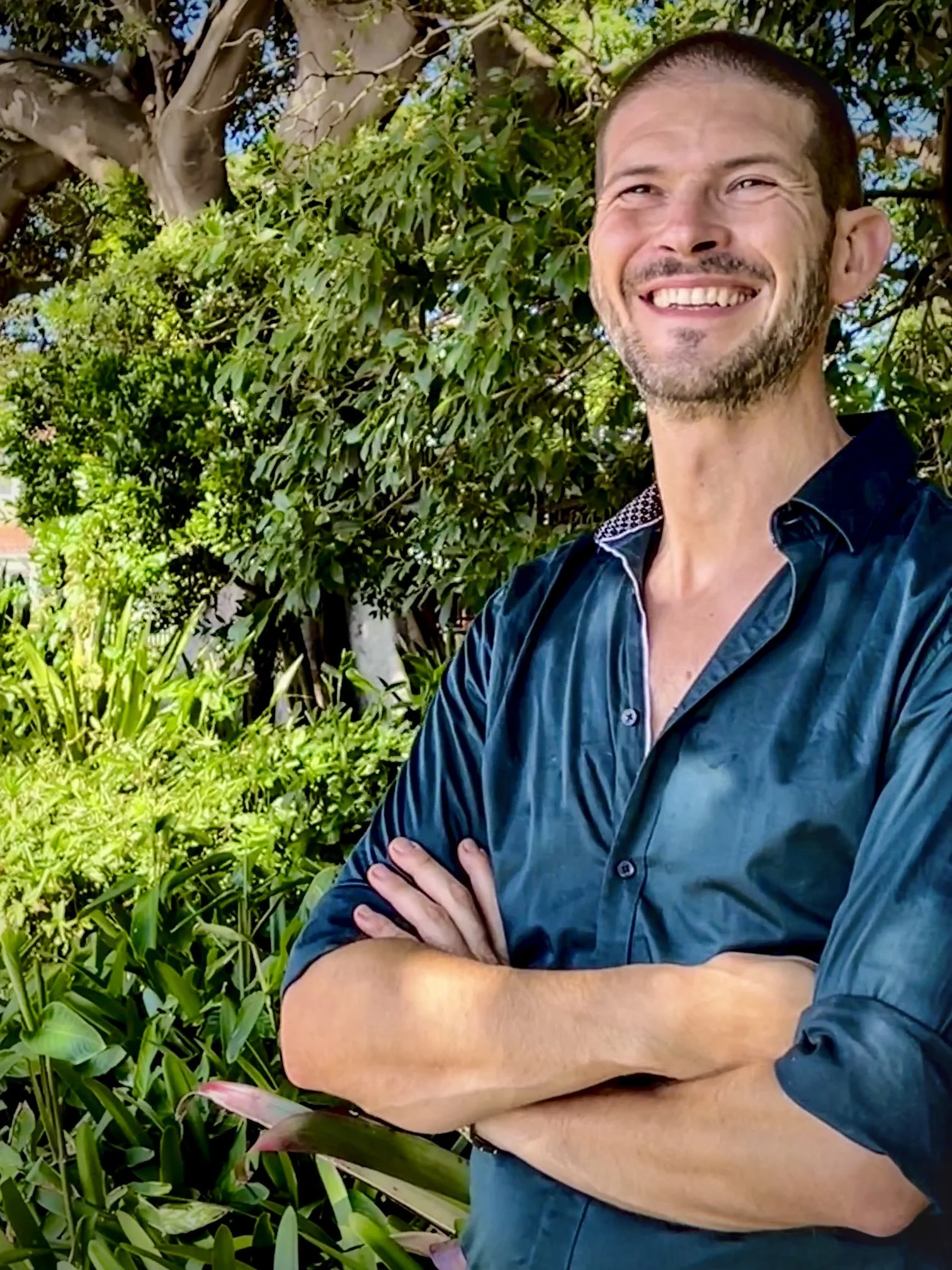 A smiling massage therapist with short hair and a beard wearing a dark shirt stands outdoors in front of lush green foliage.