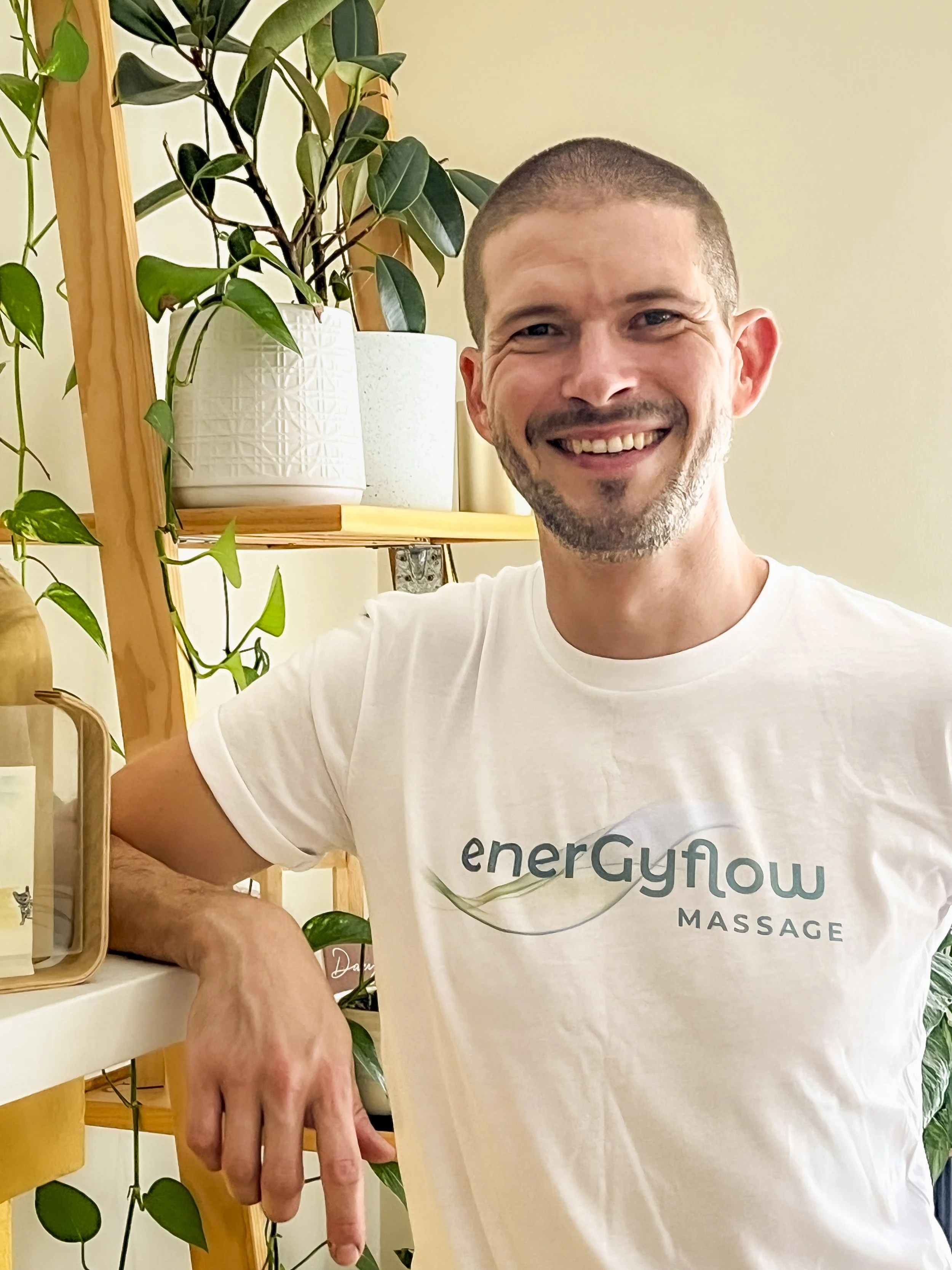 Smiling massage therapist wearing a white "enerGyflow massage" t-shirt, standing next to a wooden shelf with potted plants.