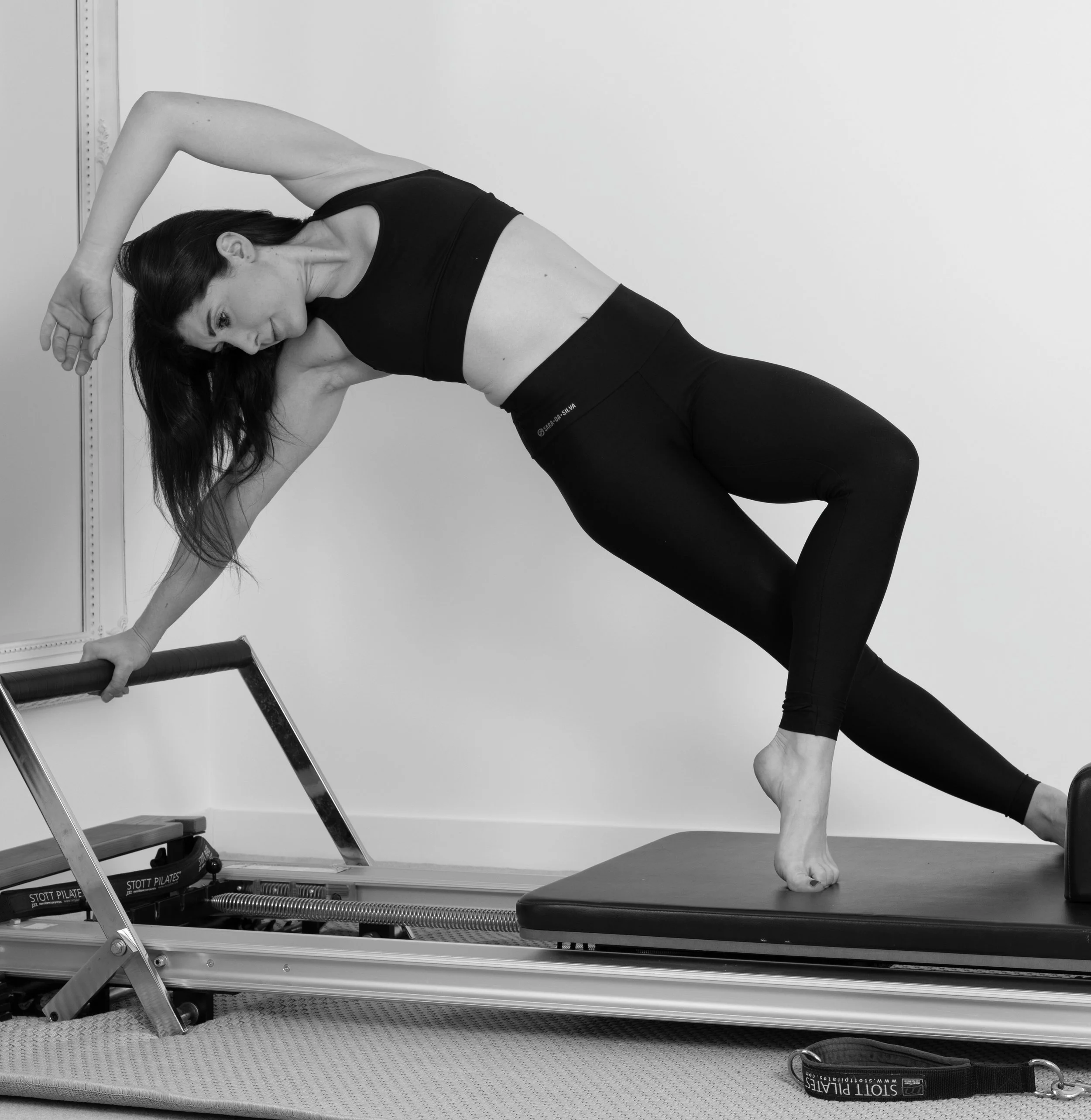 A woman performing a stretch on an Pilates reformer machine with one hand on a rail and the other hand behind her head, with one leg bent and the other extended.