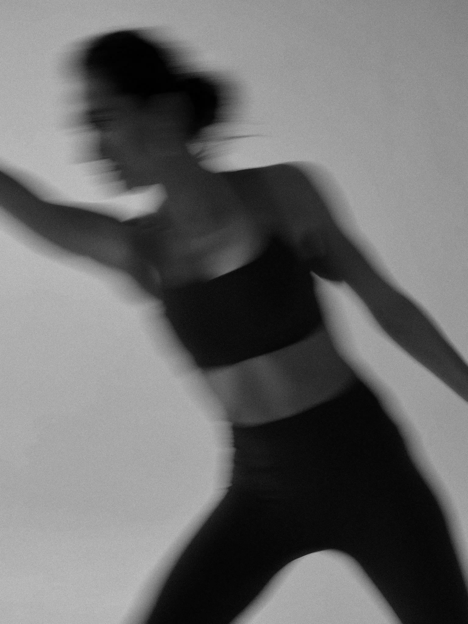 Blurred black-and-white photo of a woman in workout clothes stretching or exercising, with her arm raised and her hair tied back.