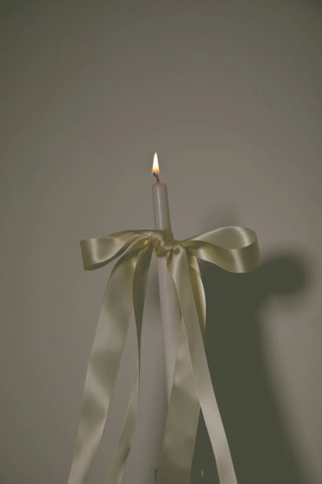 A white candle lit at the top, tied with a large satin ribbon bow, standing upright against a plain background.
