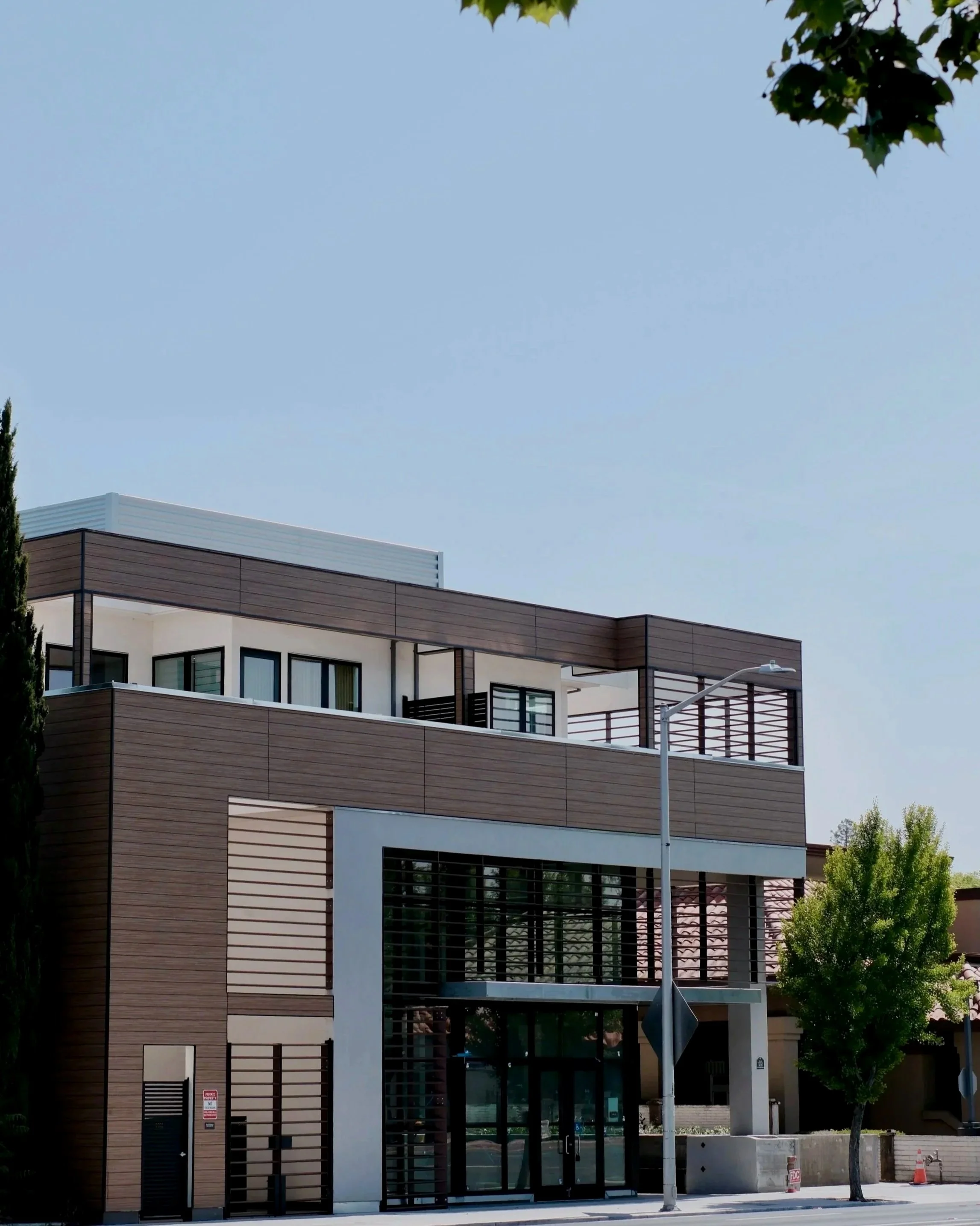 Modern multi-story building with large glass entrance, wooden panel exterior, and balcony, surrounded by trees and a clear blue sky.