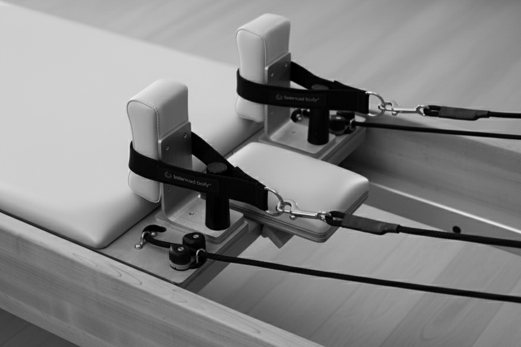 Reformer exercise machine with padded carriage, straps, and pulleys on a wooden floor.