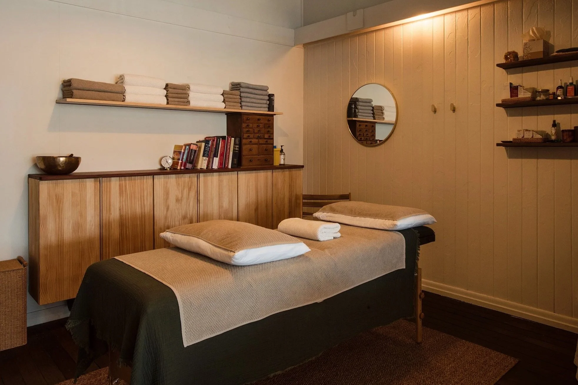 A bedroom with two beds, each covered with beige and dark bedding, and white towels placed on top. The room has wooden furniture including a cabinet with books, a small drawer chest, and a sideboard. Several folded towels are stacked on a wall shelf, and a round mirror is mounted on the wall. The wall has vertical wooden paneling, and there are shelves with various personal care items. The room has warm lighting.