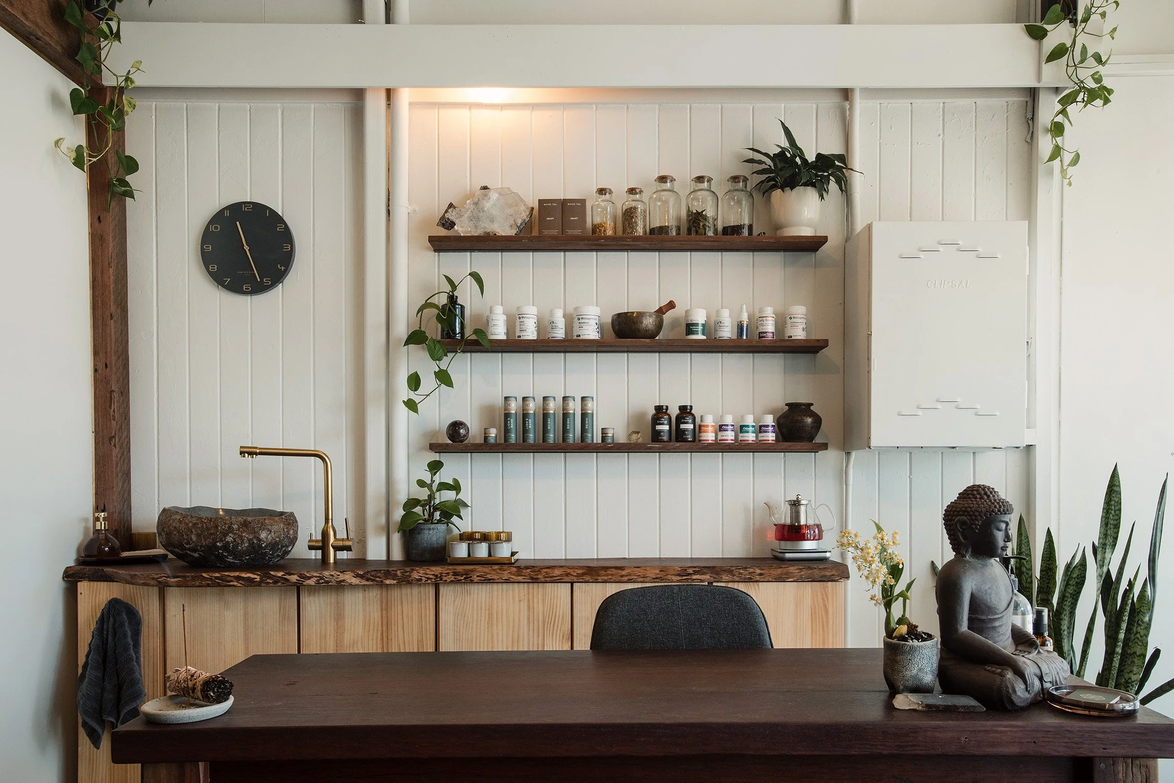 Interior of a modern, minimalist room with white paneled walls. There are three wooden shelves displaying jars, plants, and decorative items. A black wall clock is on the left side of the wall. The room features a wooden counter with a stone bowl, a golden faucet, and a small Buddha statue on the right. Various plants, bottles, and a teapot are also visible.