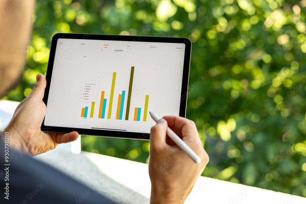 Person holding a tablet displaying a colorful bar chart outdoors with a background of green foliage.