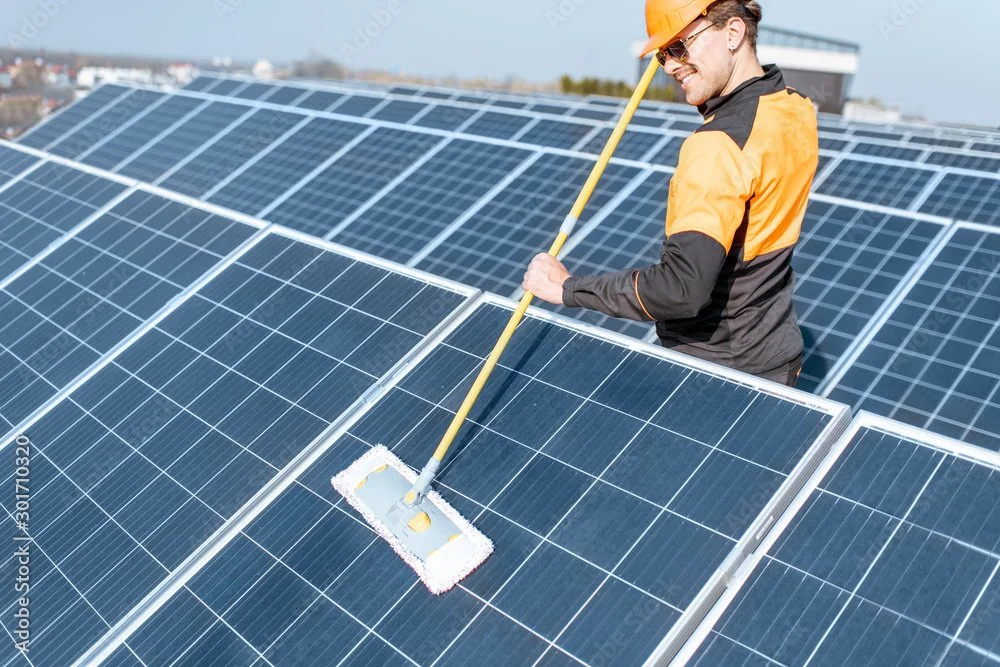 A man in safety gear cleaning a large solar panel array on a rooftop.