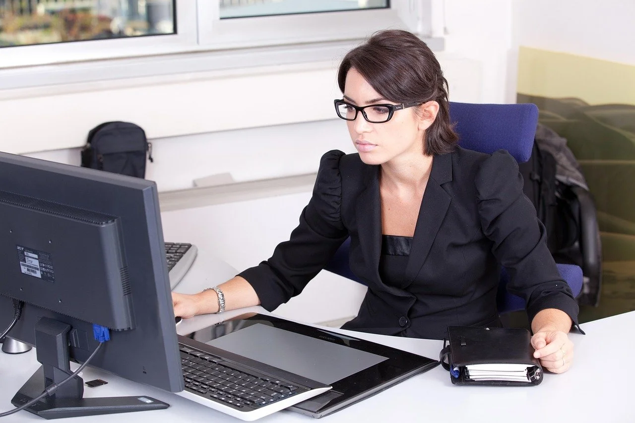 A woman with dark hair and glasses working at her desk with a computer, tablet, and notebook in an office space.