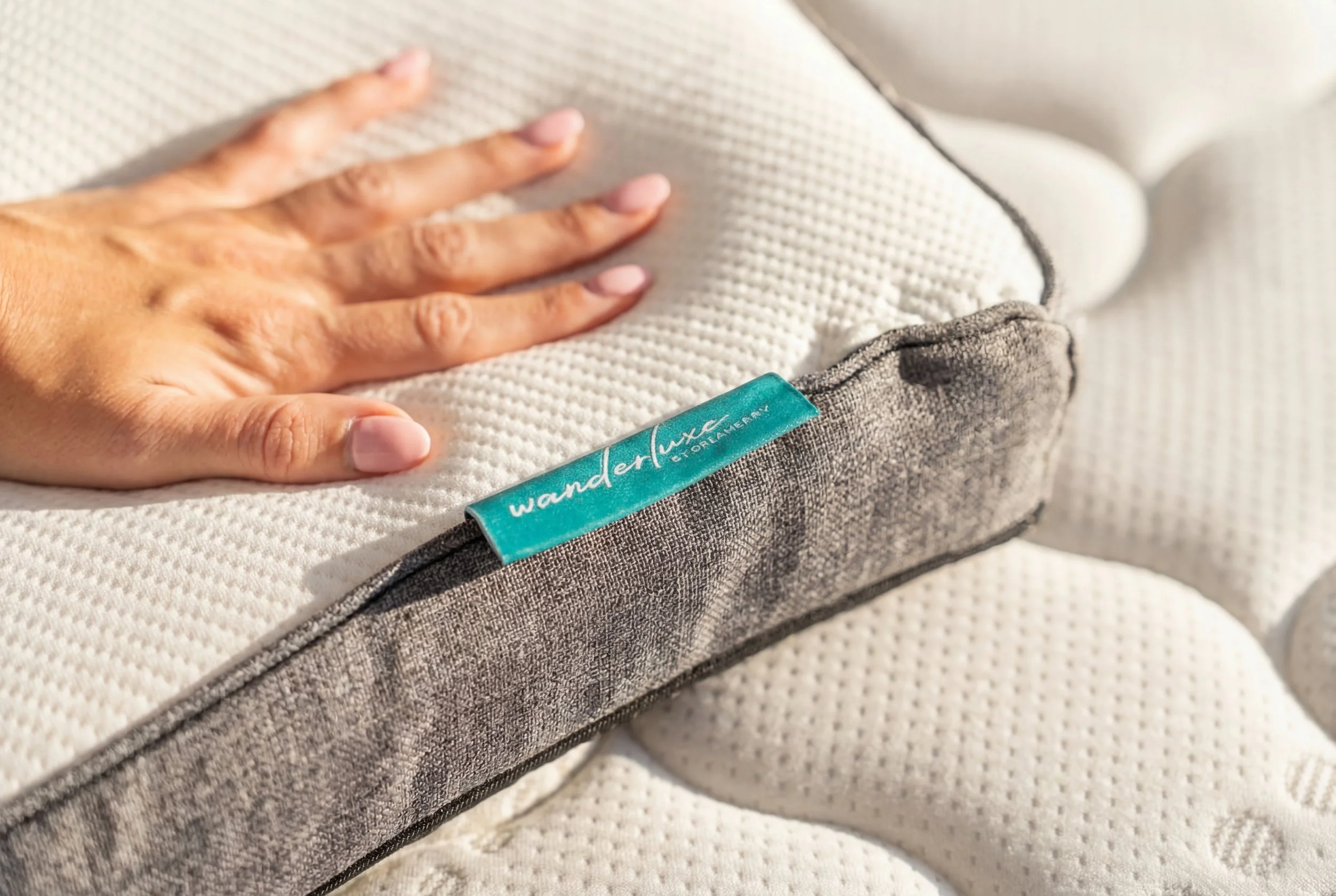 Close-up of a hand resting on a mattress with a teal tag that reads 'wandler luxe etorlamem' and a white textured and gray fabric surface.