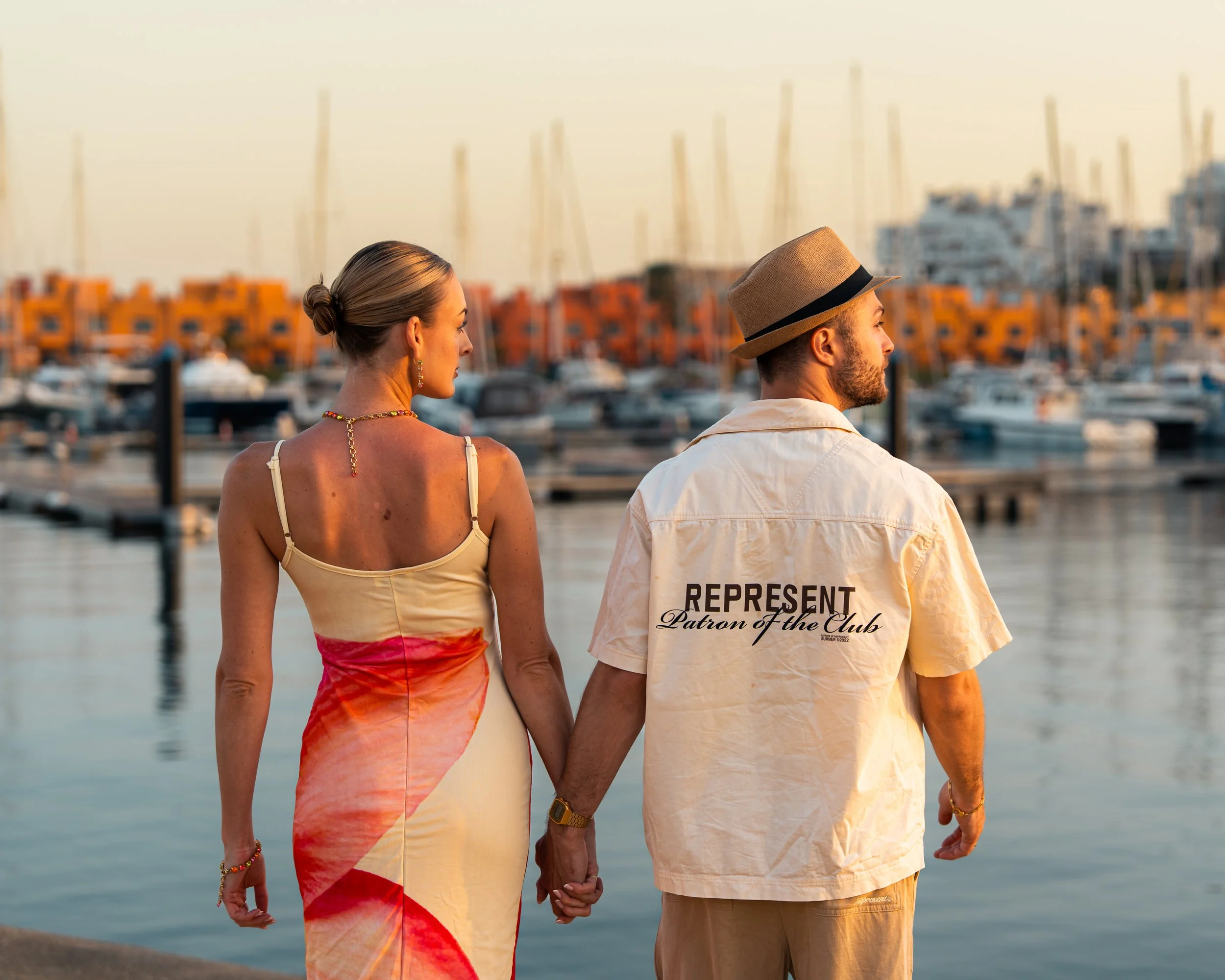 A couple holding hands by the harbor at sunset, with boats and buildings in the background.