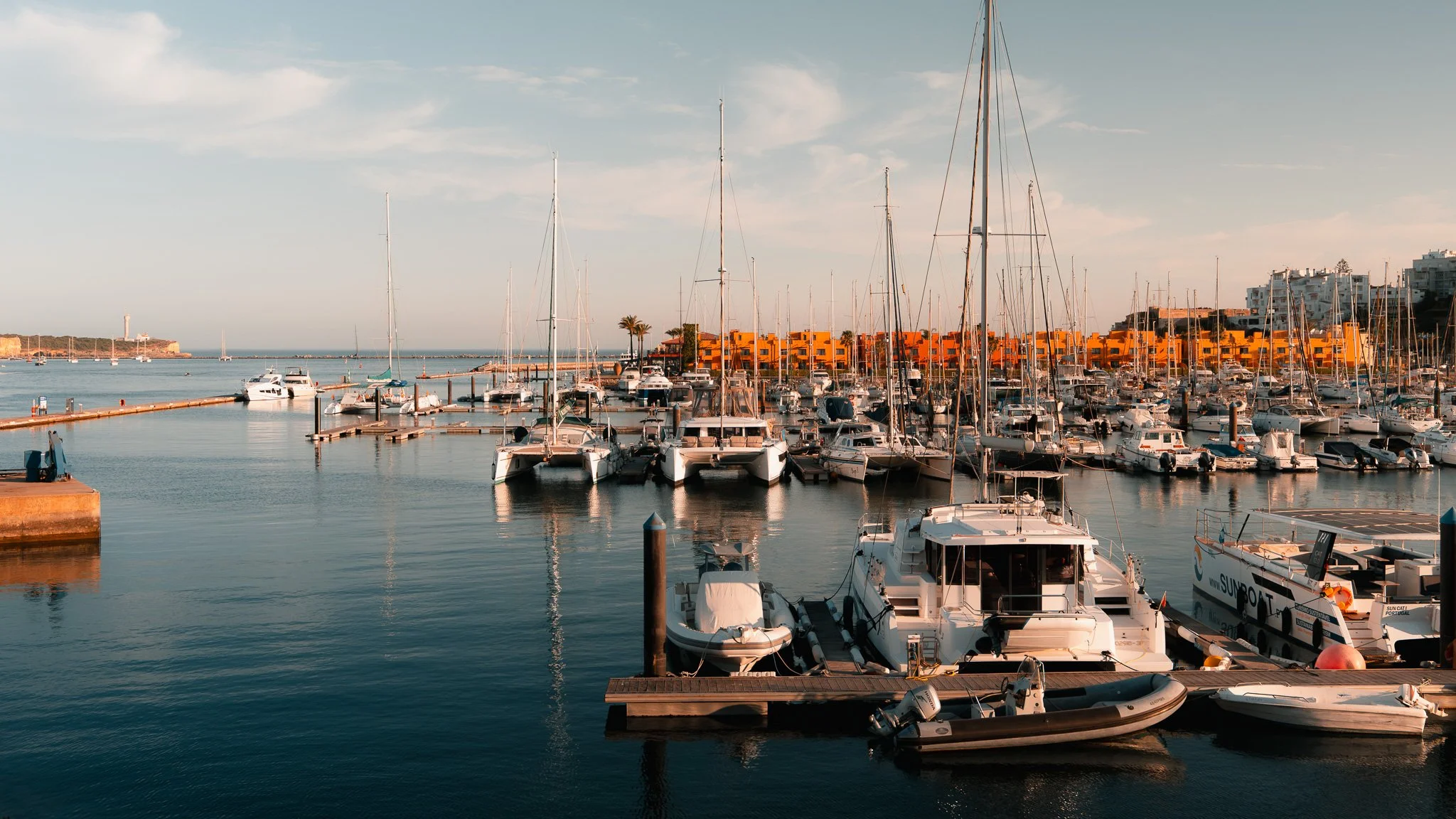 Marina filled with sailboats and yachts docked by colorful yellow buildings under a partly cloudy sky.