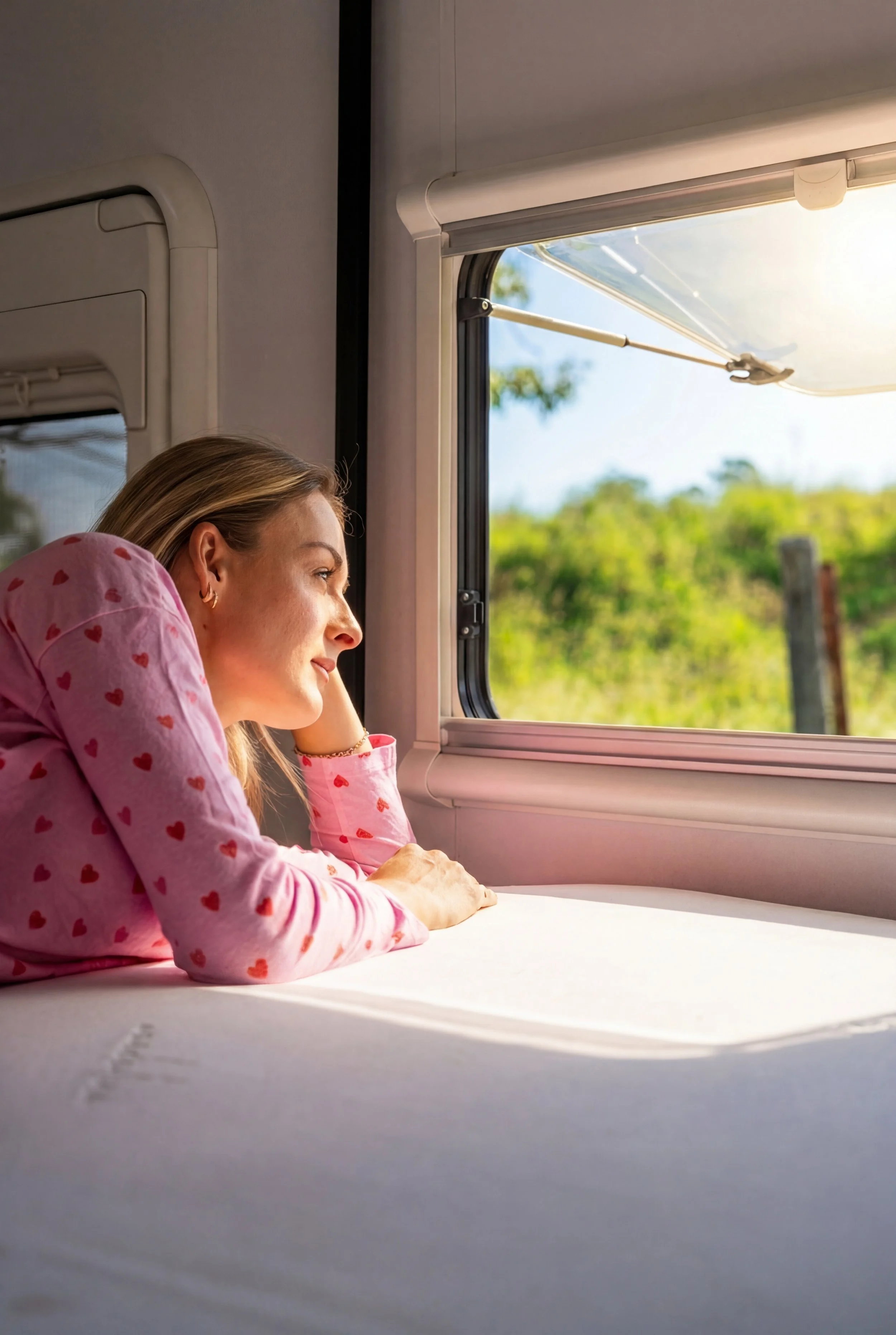 A woman in pink pajamas with red hearts is lying on a bed, gazing out a window at a sunny green landscape while resting her head on her hand.