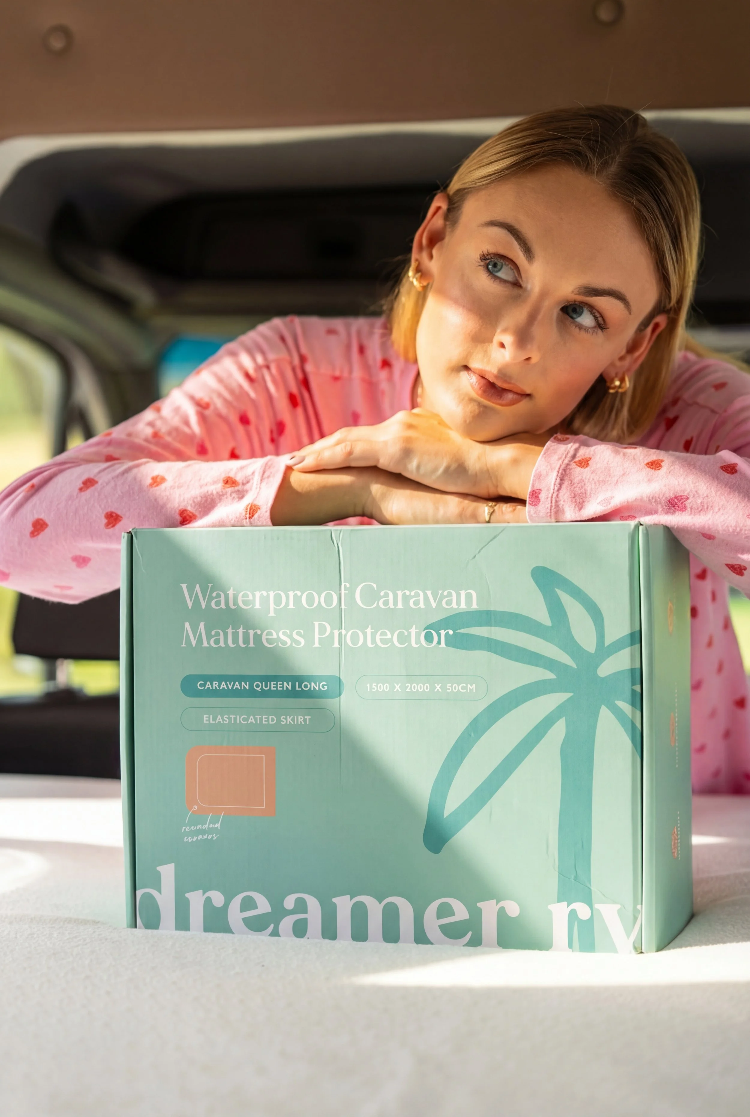A young woman with blonde hair and blue eyes resting her chin on her hands, leaning on a cardboard box labeled "Waterproof Caravan Mattress Protector" inside a vehicle. She is wearing a pink shirt with red hearts.