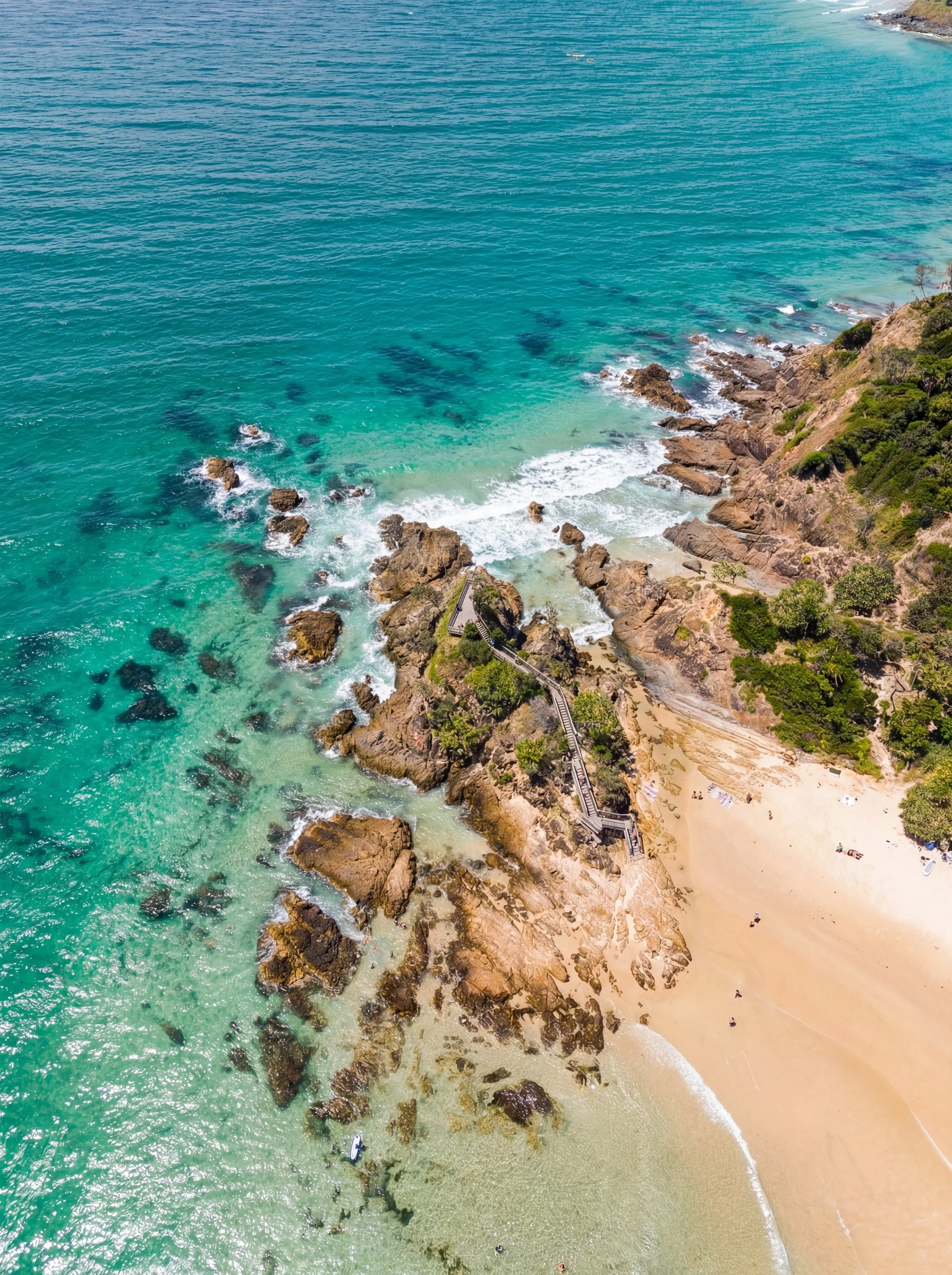 Aerial view of a beach with turquoise water, rocks, and a sandy shore. There is a staircase leading from the land down to the beach, surrounded by greenery.