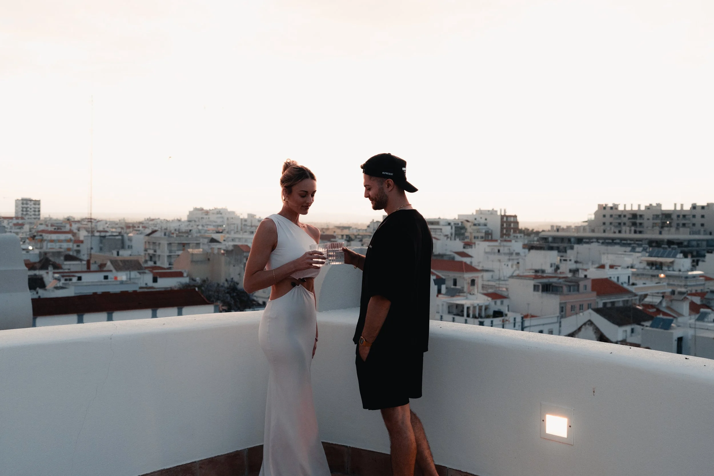 A man and woman sharing drinks on a rooftop with a cityscape in the background at sunset.