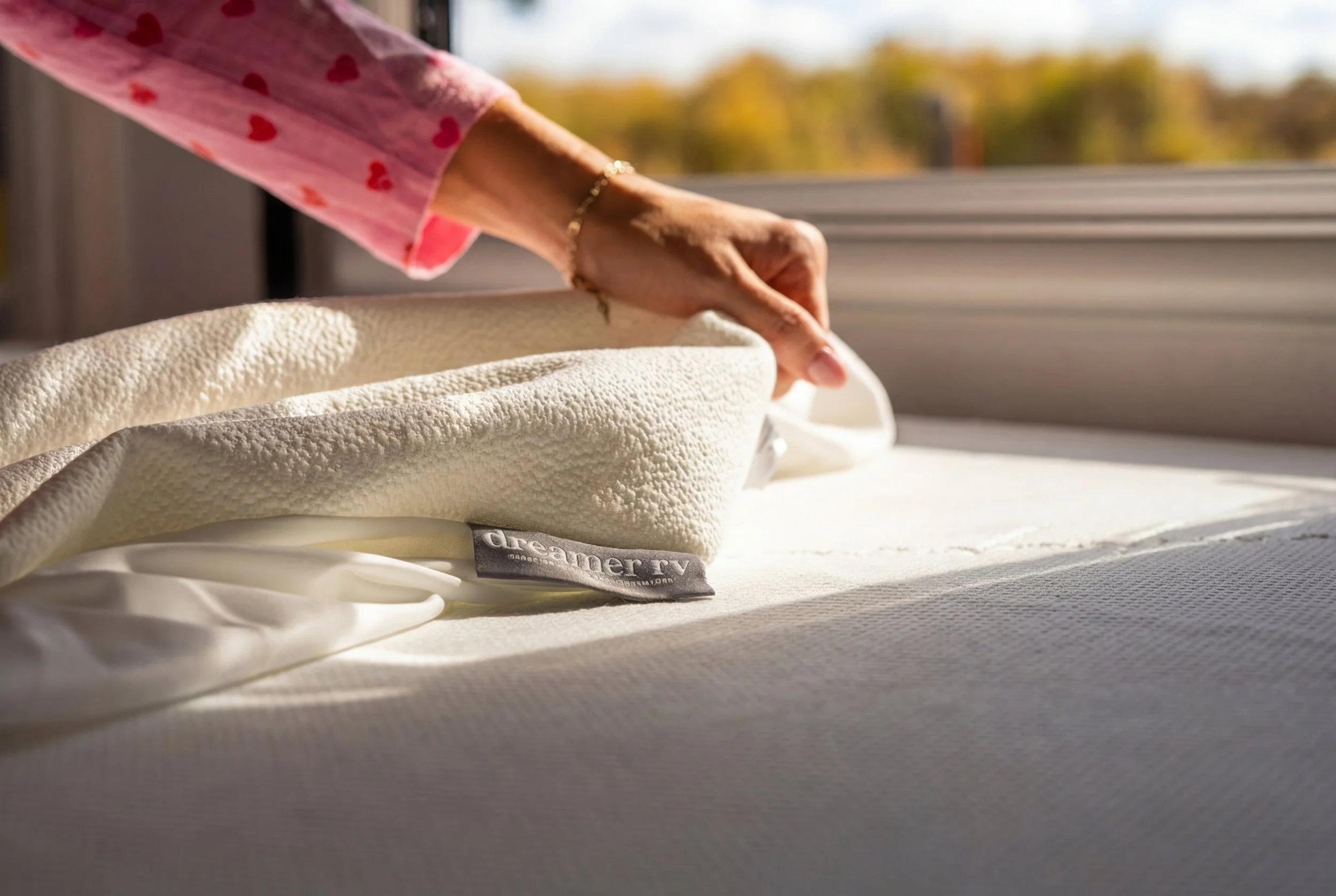 Close-up of a person's hand making a bed, with a foam mattress topper labeled 'dreamer rv,' and a window with sunlight and trees outside in the background.
