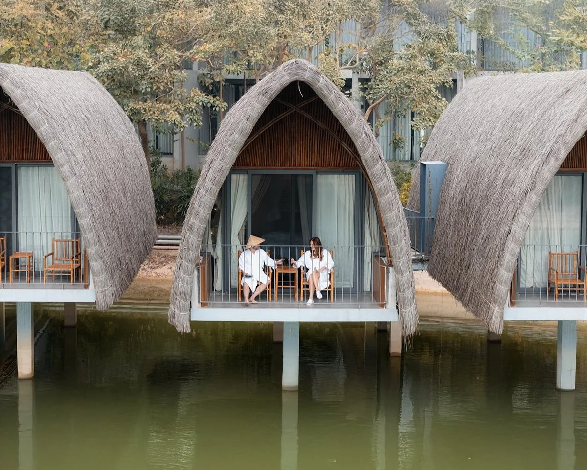 Aerial view of two stilted bungalow-style overwater accommodations with thatched roofs, featuring two women in white robes sitting at a table and talking on a balcony overlooking a body of water, surrounded by trees.