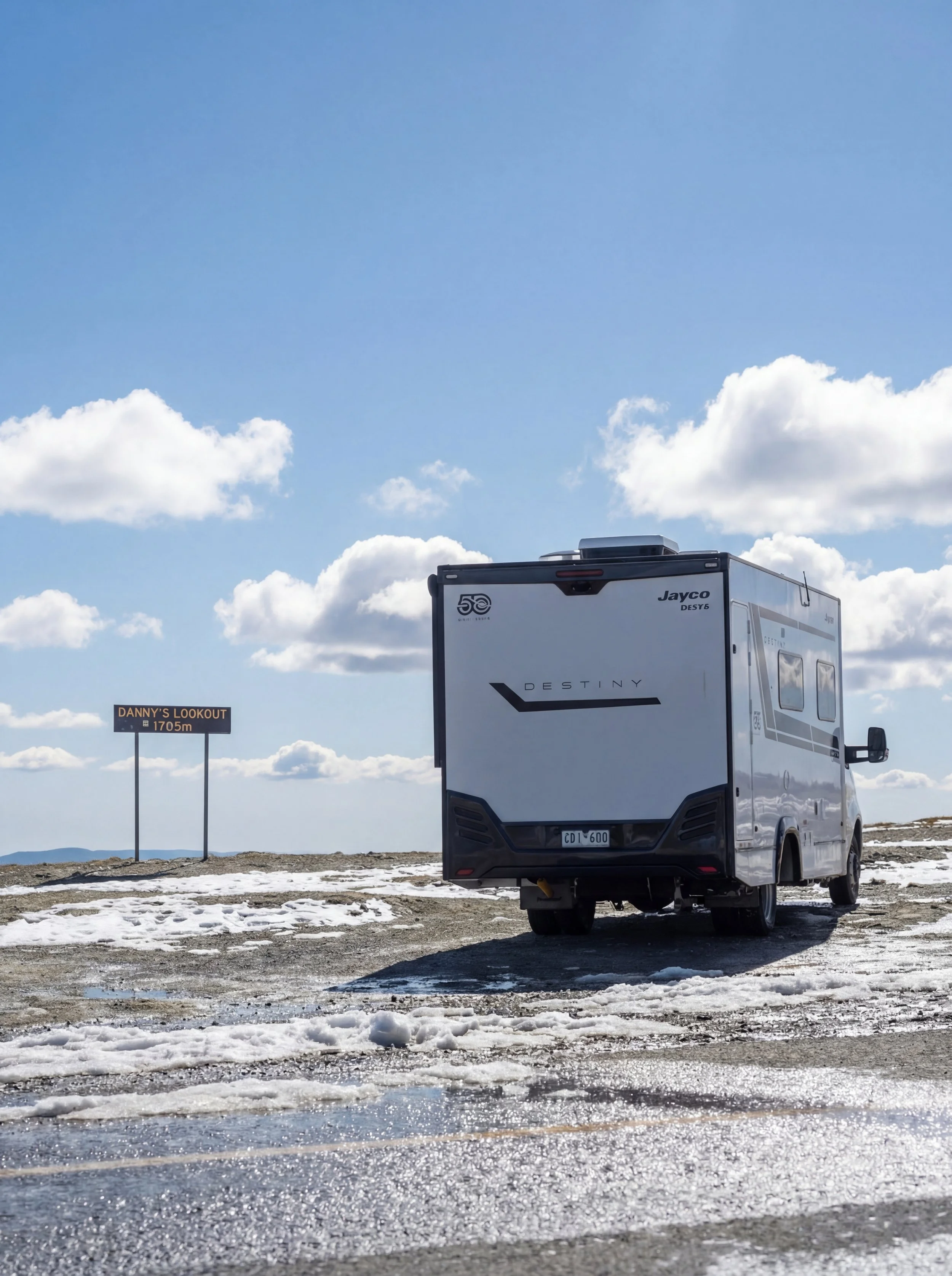A white camper van parked on a snowy, icy landscape with a blue sky and scattered clouds, and a sign that reads "DANNY'S LOOKOUT 1705m" in the background.