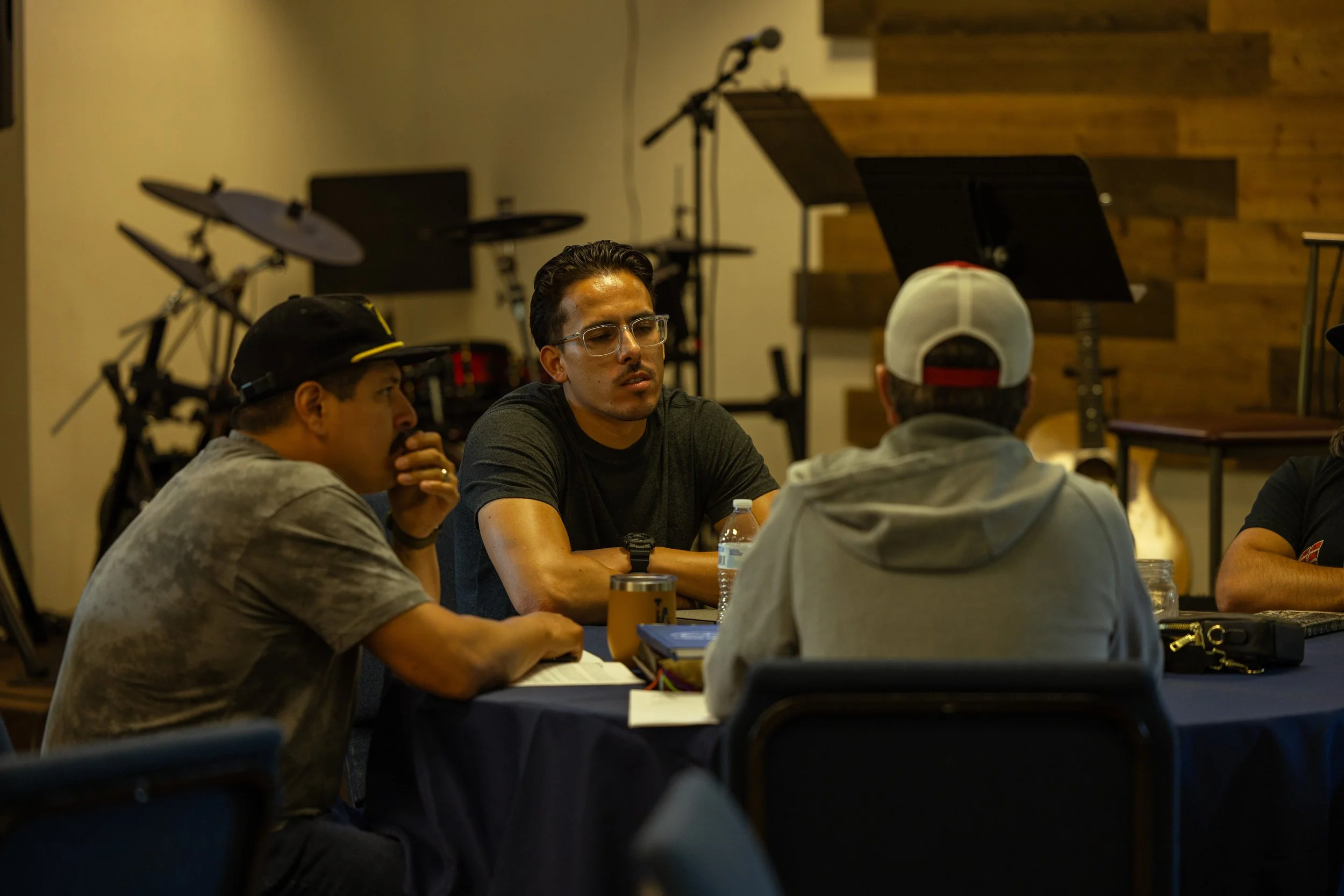 Three men sitting at a table in a room with musical instruments in the background, engaged in conversation.