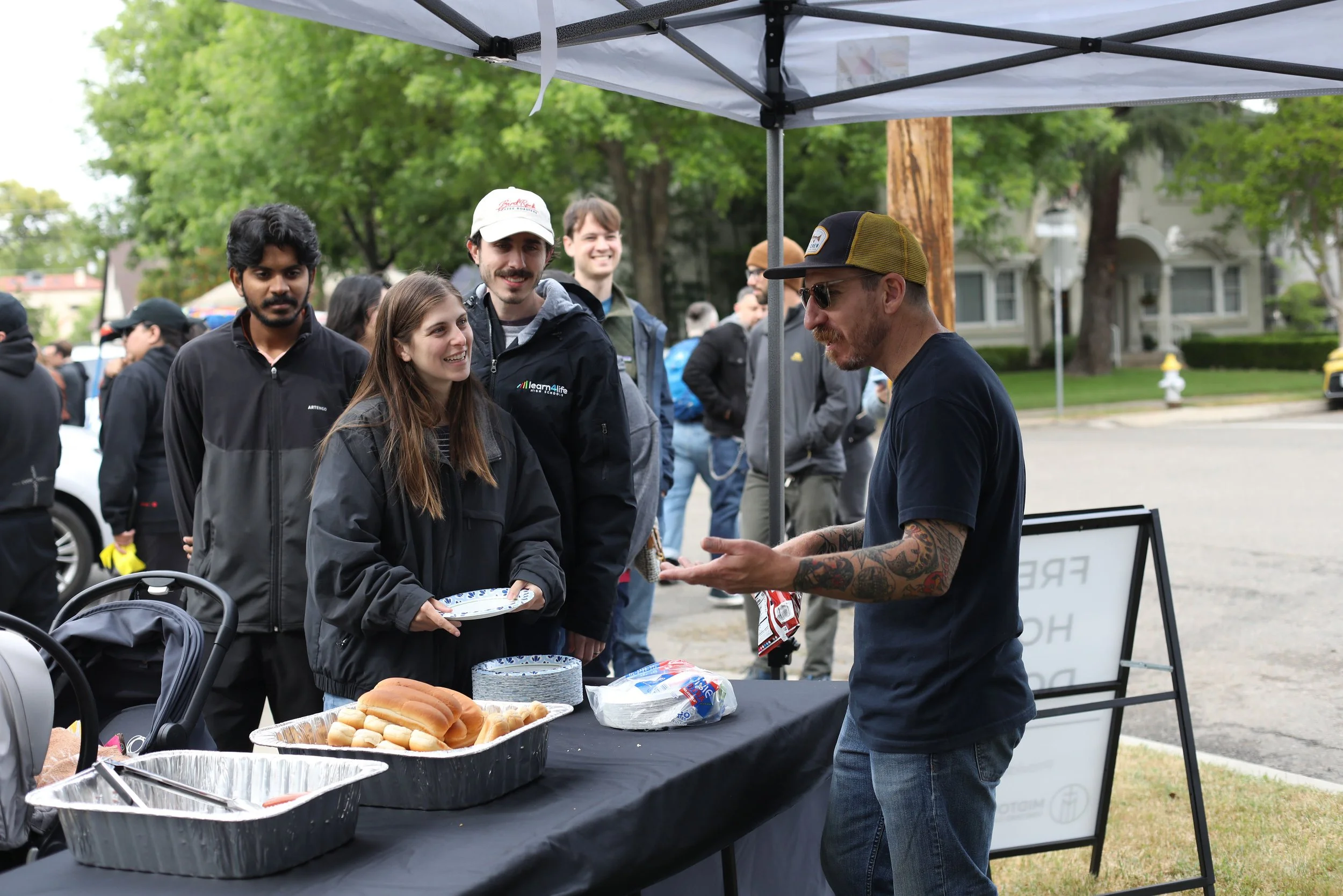 A group of young people standing in line at an outdoor food stand with food items on the table, while a man with tattoos and sunglasses talks to them. There are green trees and other people in the background.
