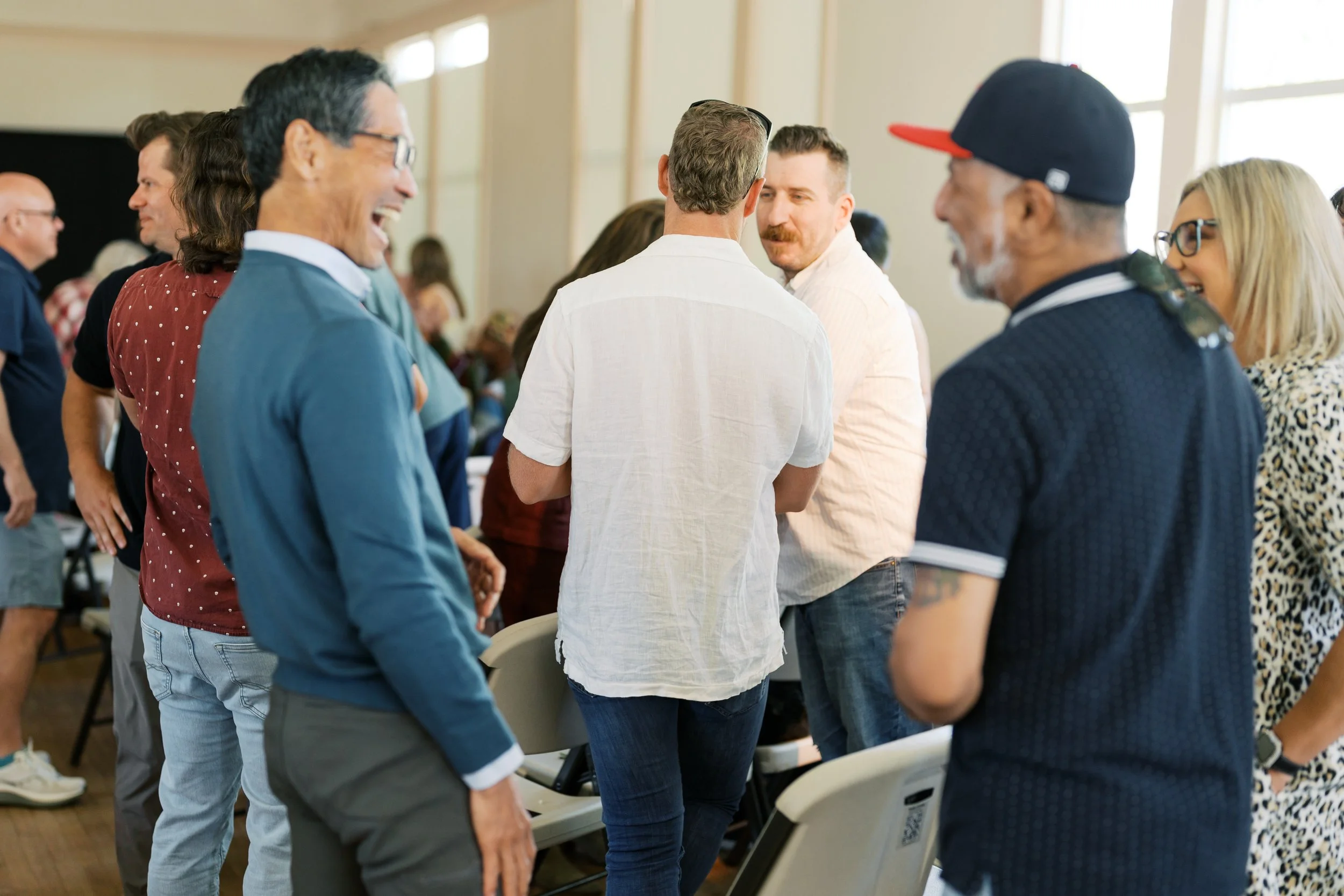 A group of people standing and facing each other in a circle at an indoor event, smiling and engaging in conversation.