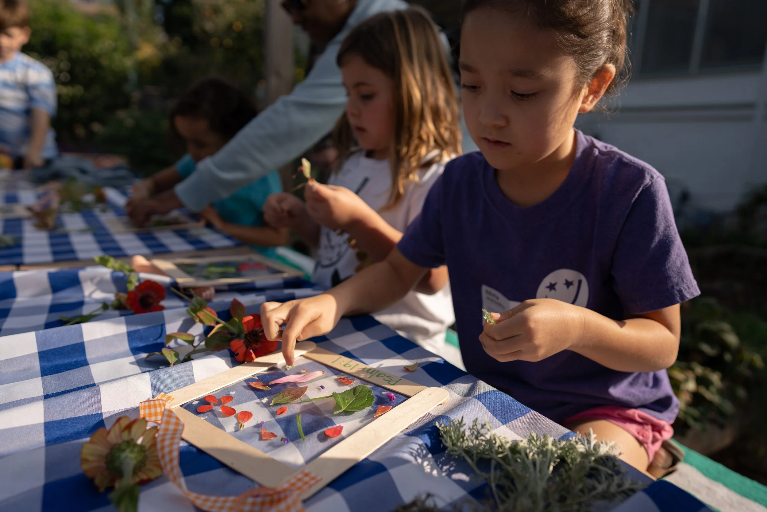 Children and adults sitting at a table outdoors, engaging in a craft activity with flowers and leaves, under a blue and white checkered tablecloth.