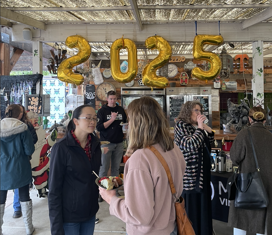 People celebrating 2023 with large gold balloons in a rustic indoor space. Several individuals are socializing, eating, and drinking. A bar setup with decorations and a Christmas theme is visible.