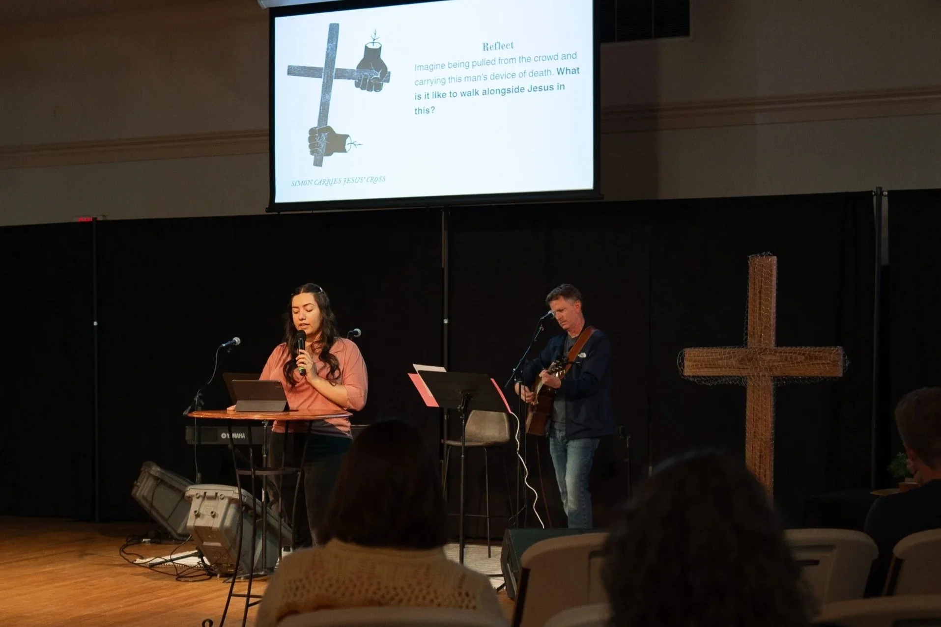 A woman and a man are on a stage in a church, performing music. The woman is singing into a microphone and playing a keyboard. The man is playing a guitar. There is a large wooden cross on the right side of the stage. Above them, a screen displays a slide with an illustration of a cross and a quote about Jesus carrying a man's device of death.