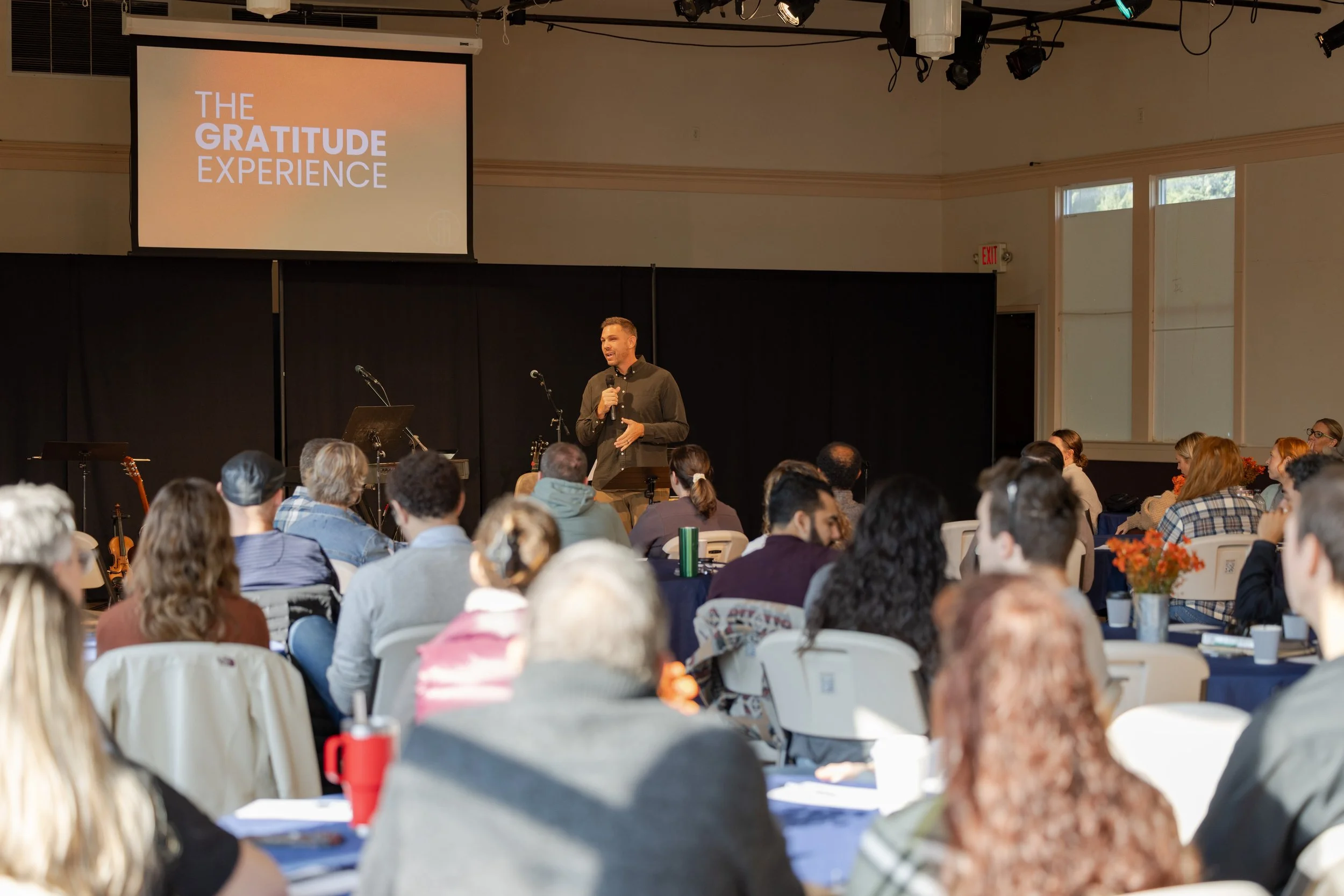 A speaker presenting on stage at an event called 'The Gratitude Experience', with an audience seated in front, including people with diverse hair types and colors. The presentation slide displays the event's name.